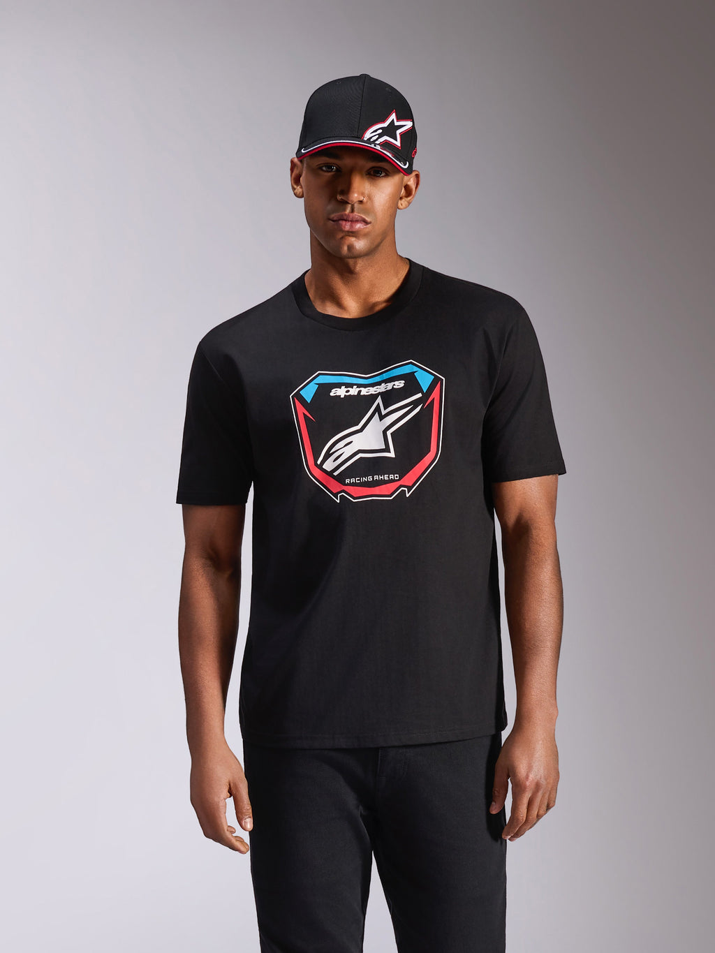 T-shirt Alpinestars Plate CSF, maglietta a maniche corte, nero, con grafica frontale ispirata alle tabelle MX con logo bianco e accenti rossi e blu, indossata da un modello con cappellino nero coordinato, streetwear casual motorsport