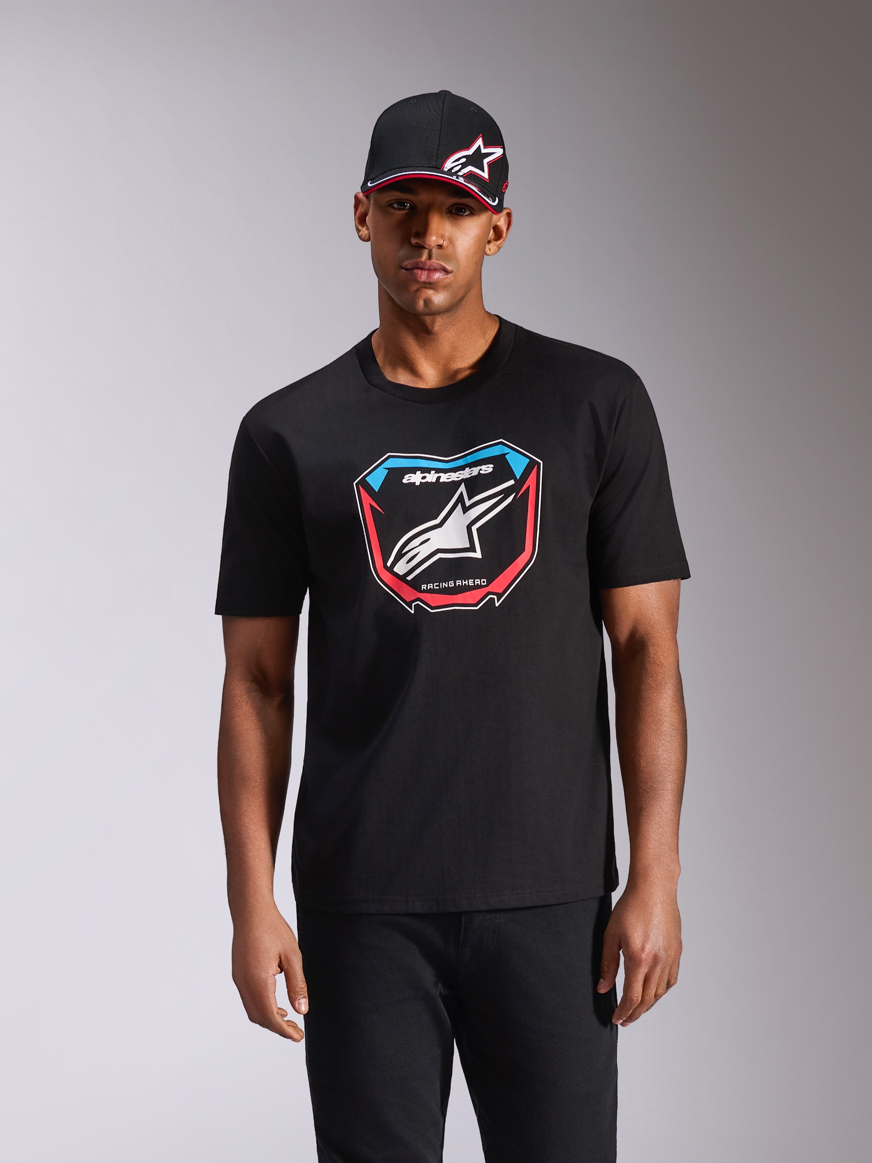 T-shirt Alpinestars Plate CSF, maglietta a maniche corte, nero, con grafica frontale ispirata alle tabelle MX con logo bianco e accenti rossi e blu, indossata da un modello con cappellino nero coordinato, streetwear casual motorsport