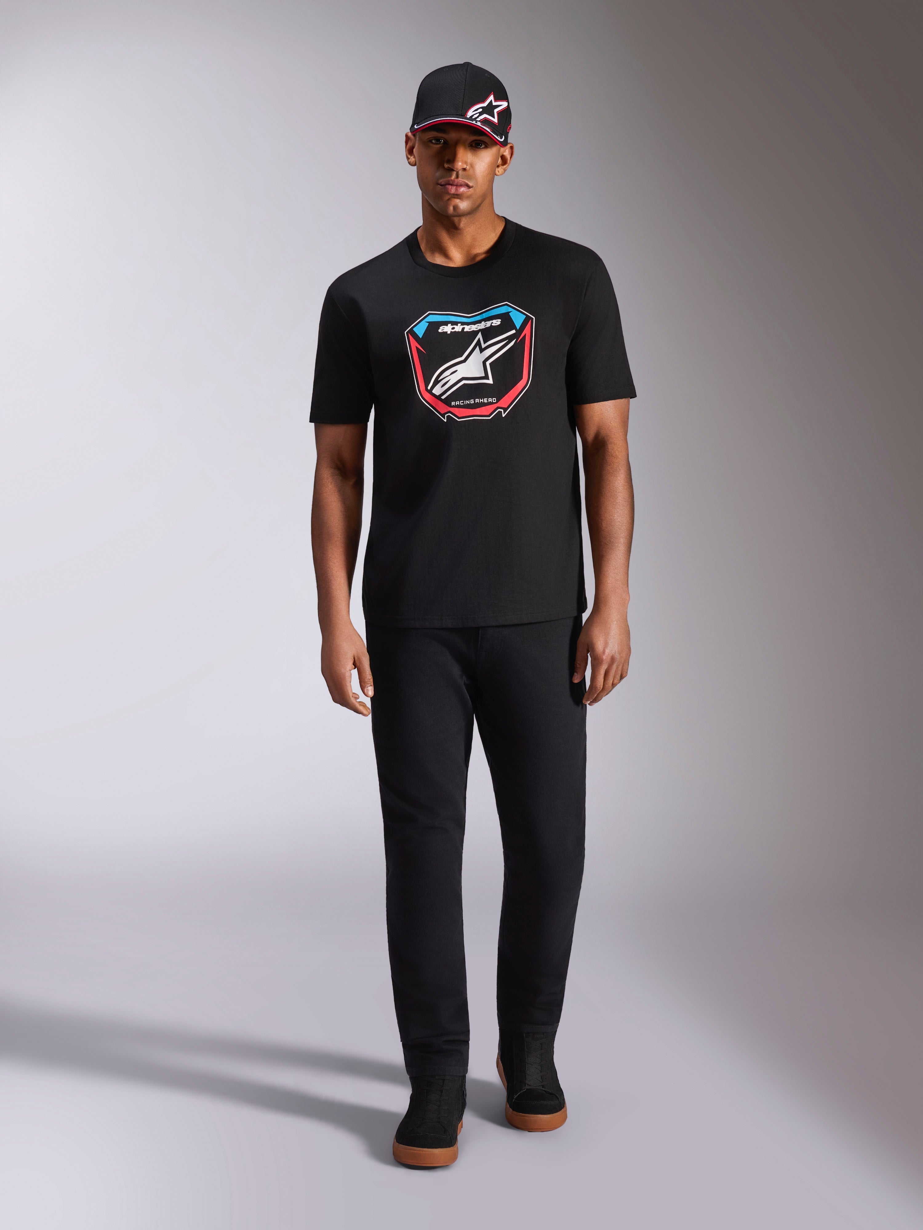 Alpinestars Plate CSF Tee, maglietta a maniche corte, nero, grafica frontale con placca MX, logo Astars bianco e accenti blu e rossi, indossata da un modello con cappellino e pantaloni neri coordinati, abbigliamento sportivo casual motorsport