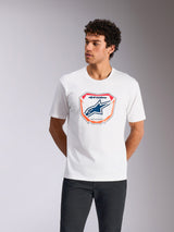 Alpinestars Plate CSF Tee, maglietta a maniche corte, bianco, con grafica frontale a placca Astars MX con logo blu e bordo arancione, indossata da un modello, streetwear casual motorsport