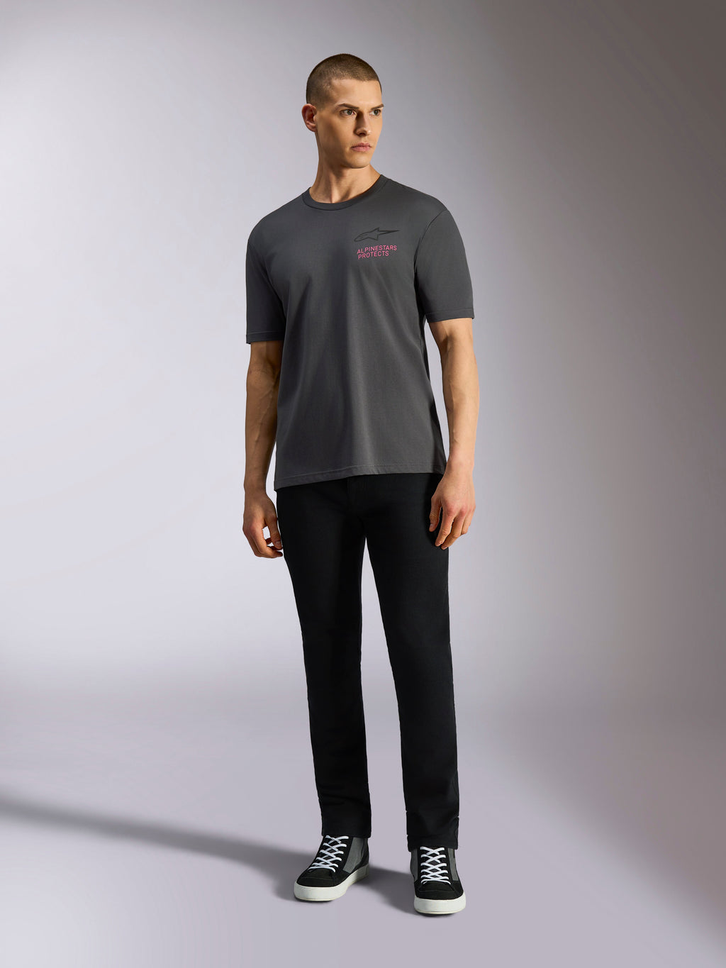 Alpinestars Pop Helmet CSF Tee, t-shirt a maniche corte, grigio antracite, con logo Alpinestars rosa sul petto, indossata da un modello uomo con pantaloni neri e sneaker, abbigliamento streetwear casual motorsport