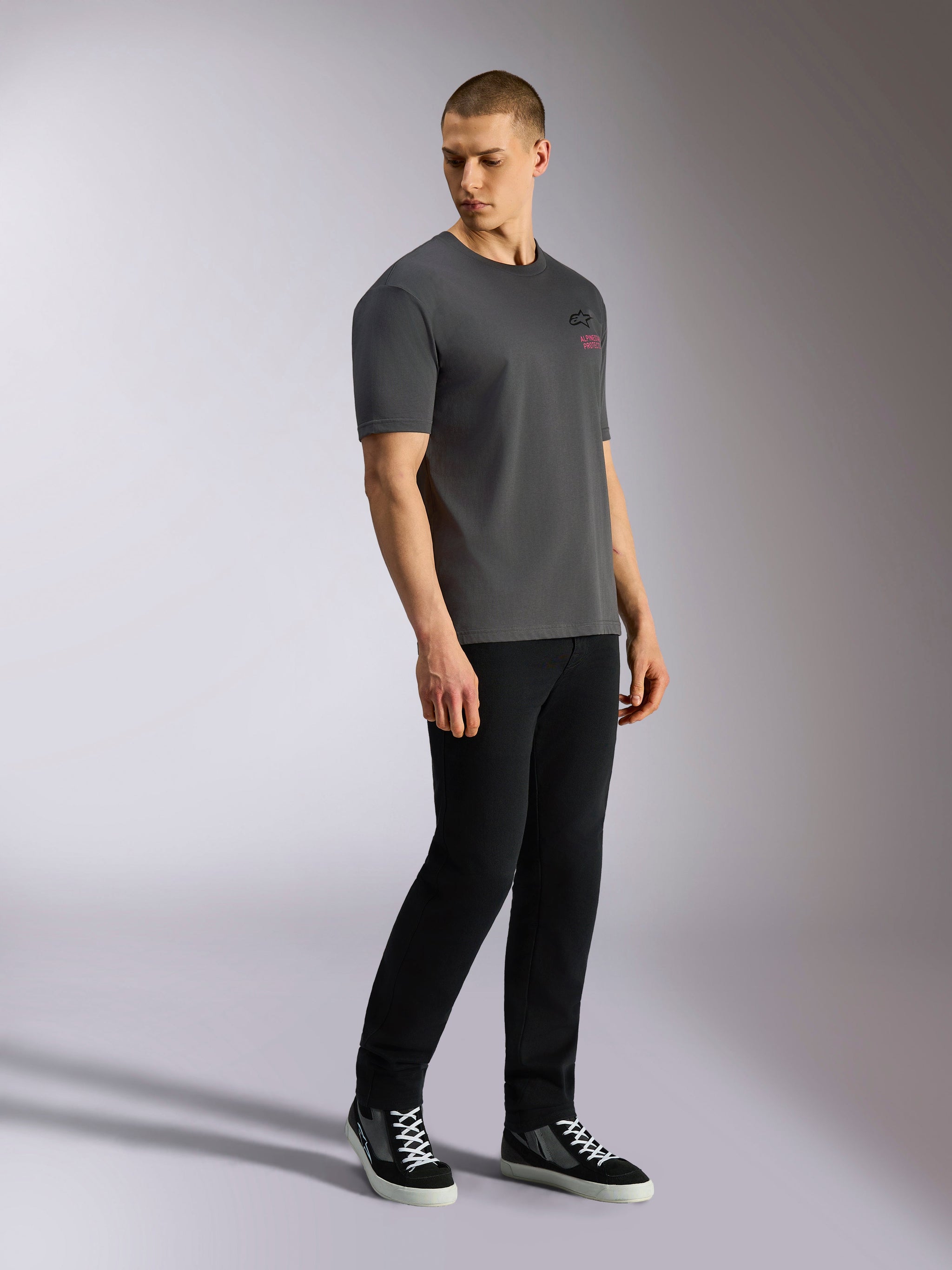 Alpinestars Pop Helmet CSF Tee, t-shirt a maniche corte, grigio antracite, indossata da un modello con pantaloni neri e sneakers, con logo Astars nero frontale e scritte rosa, streetwear casual ispirato al motorsport