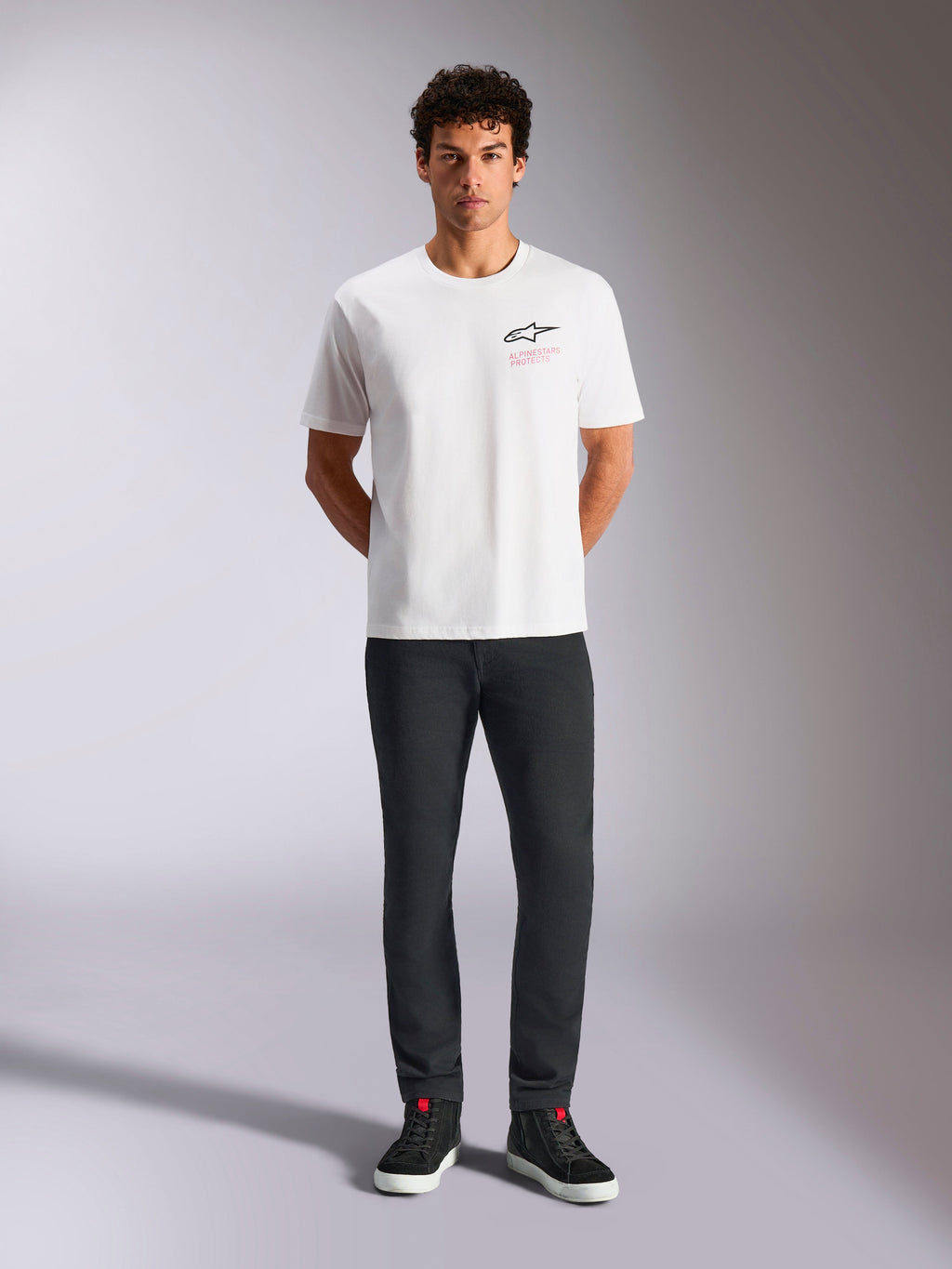 Maglietta Alpinestars Pop Helmet CSF, T-shirt a maniche corte, bianca, indossata da un modello con pantaloni neri e sneakers alte nere, logo Astars frontale con scritta rosa 'Alpinestars Protects', streetwear casual motorsport
