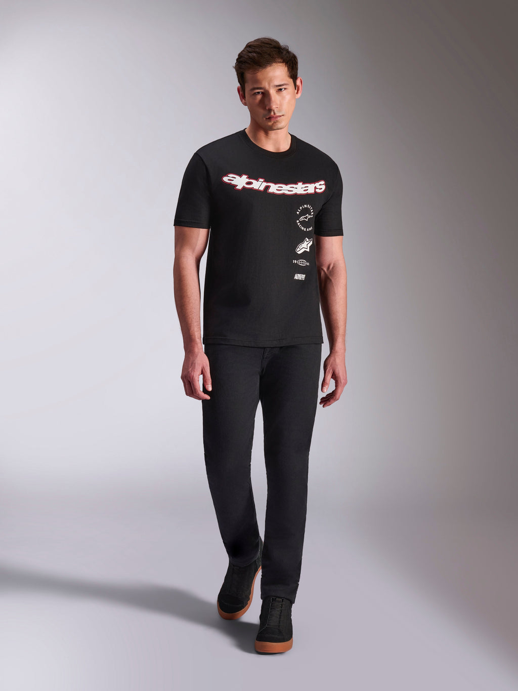 Alpinestars Abus CSF Tee, t-shirt a maniche corte, nera, logo curvo rosso e bianco con loghi multipli sovrapposti, indossata da un modello con pantaloni neri e sneakers nere, streetwear casual motorsport