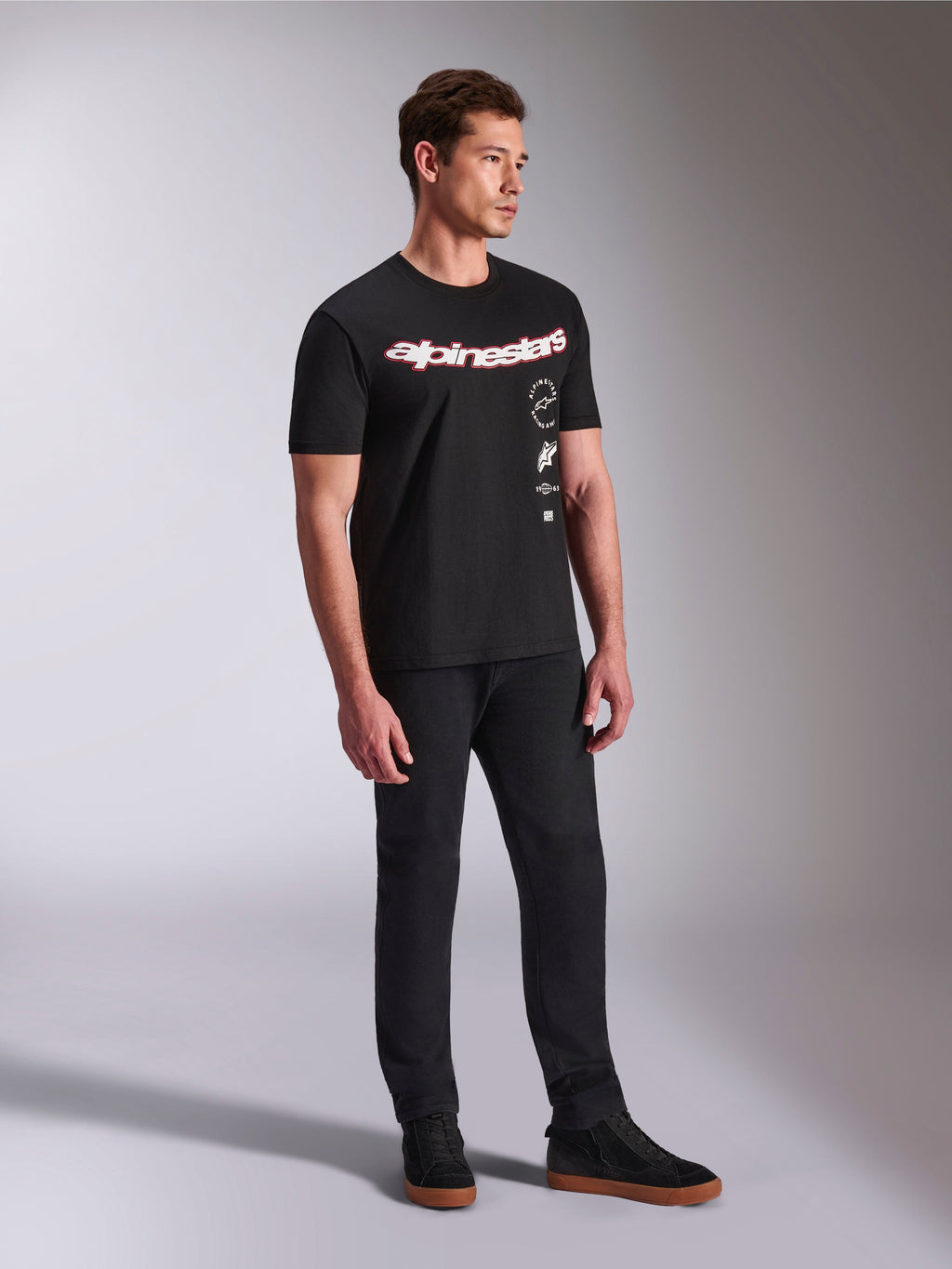 T-shirt Alpinestars Abus CSF, maglietta a maniche corte, nera, con logo Alpinestars curvo rosso e bianco e grafiche verticali impilate, indossata con pantaloni neri e sneaker, streetwear casual ispirato al motorsport