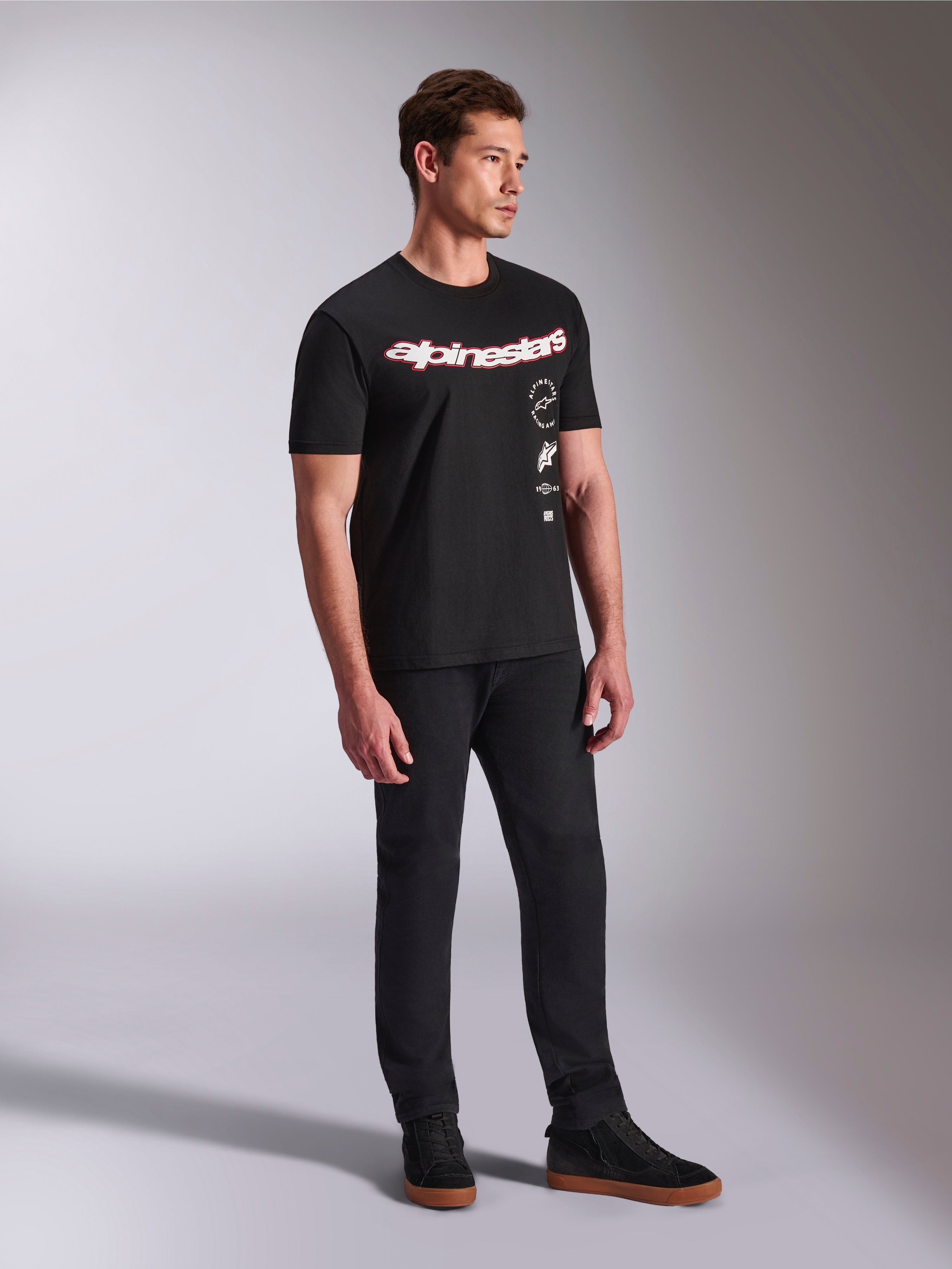 T-shirt Alpinestars Abus CSF, maglietta a maniche corte, nera, con logo Alpinestars curvo rosso e bianco e grafiche verticali impilate, indossata con pantaloni neri e sneaker, streetwear casual ispirato al motorsport
