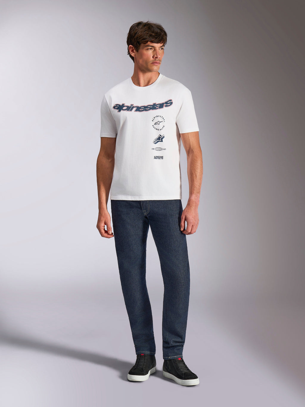 Alpinestars Abus CSF Tee, t-shirt a maniche corte, bianca, con logo Alpinestars curvo e grafiche multiple sul petto, indossata da un modello con jeans blu scuro e sneaker nere, streetwear casual ispirato al motorsport