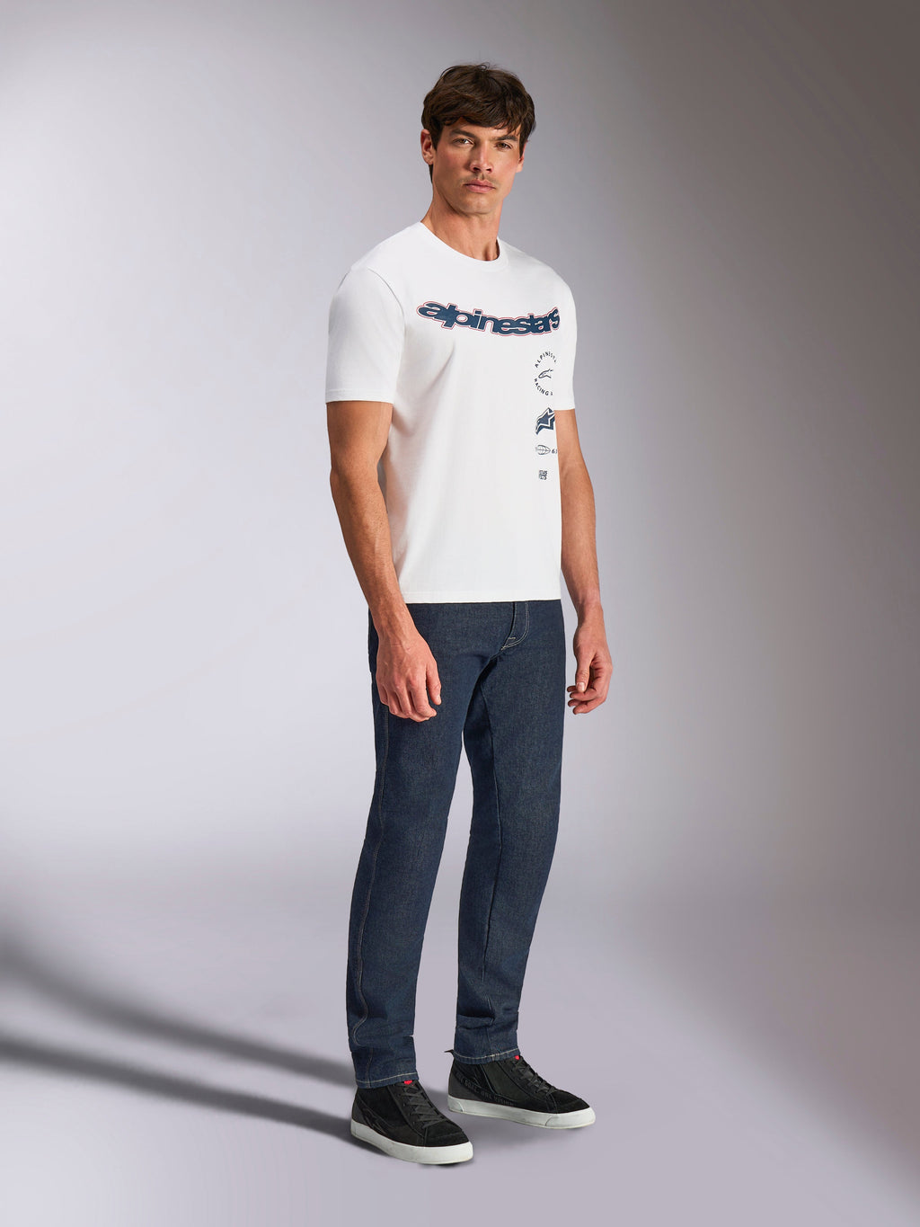 Alpinestars Abus CSF Tee, t-shirt a maniche corte, bianca, design con logo curvo Alpinestars e loghi impilati multipli, indossata da un modello maschile con jeans blu scuro e sneakers nere, streetwear casual ispirato al motorsport