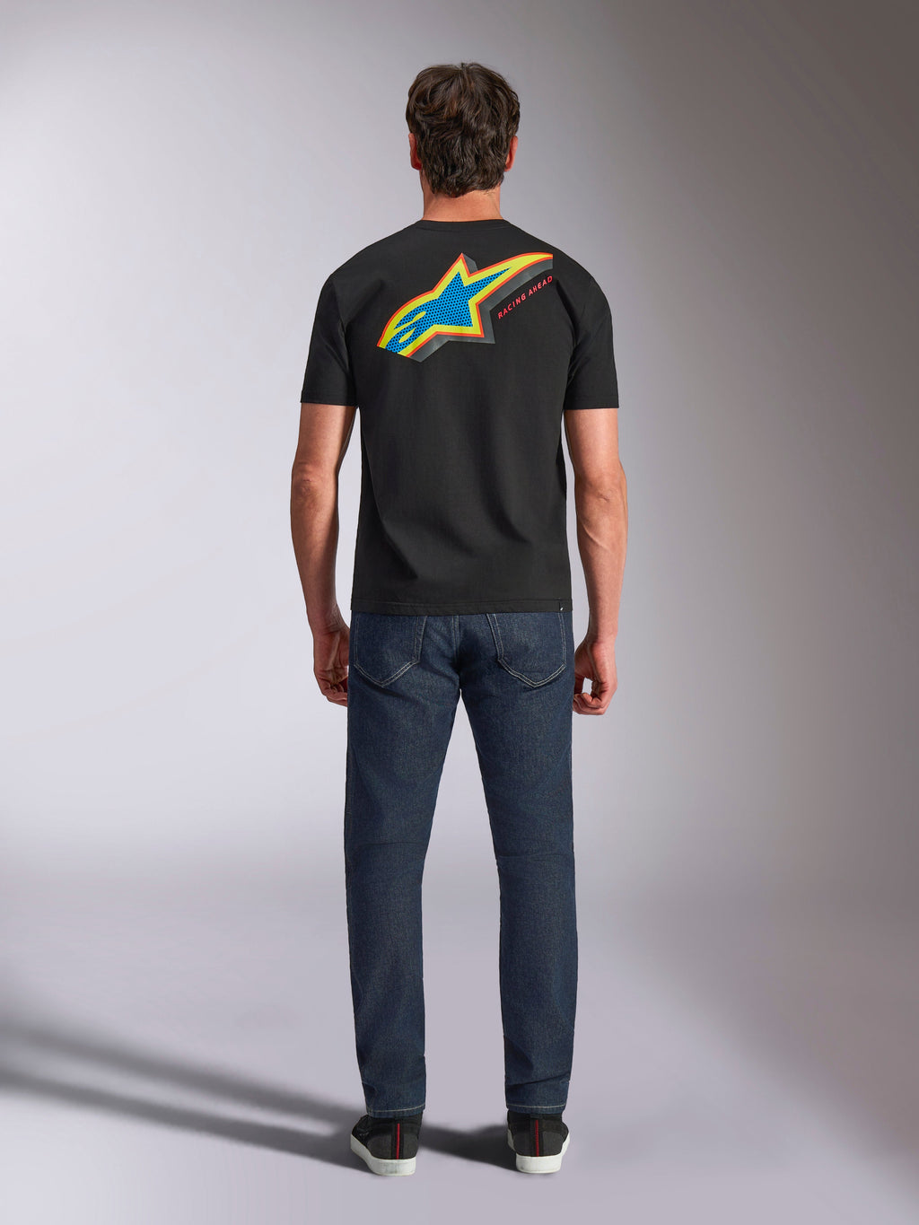Alpinestars Angler CSF Tee, t-shirt a maniche corte, nero, vista posteriore del modello con grande logo Astars a stella multicolore e scritta 'RACING AHEAD', abbinata a jeans blu scuro, streetwear casual motorsport
