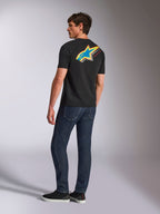 Alpinestars Angler CSF Tee, maglietta a maniche corte, nero, con un grande logo Astars multicolore sul retro con dettagli traforati in blu, giallo e rosso, indossata con jeans blu scuro, streetwear casual ispirato al motorsport