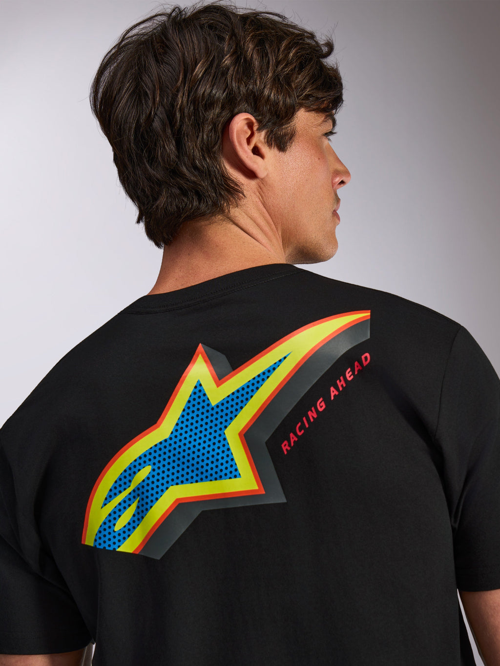 Alpinestars Angler CSF Tee, t-shirt a maniche corte nera, grande logo Astars multicolore sulla schiena con riempimento blu traforato, contorni gialli e rossi e scritta 'RACING AHEAD', abbigliamento casual streetwear motorsport