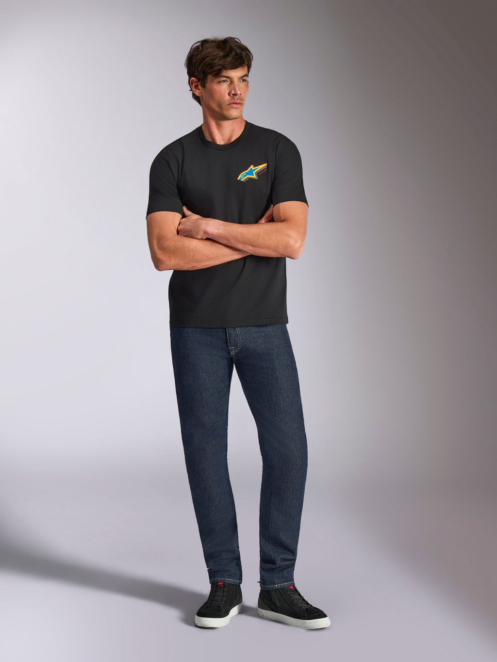 Alpinestars Angler CSF Tee, maglietta a maniche corte, nera, vivace logo Astars multicolore sul petto, indossata da un modello con le braccia incrociate, jeans in denim scuro e sneakers nere, streetwear casual motorsport
