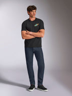 Alpinestars Angler CSF Tee, maglietta a maniche corte, nera, vivace logo Astars multicolore sul petto, indossata da un modello con le braccia incrociate, jeans in denim scuro e sneakers nere, streetwear casual motorsport