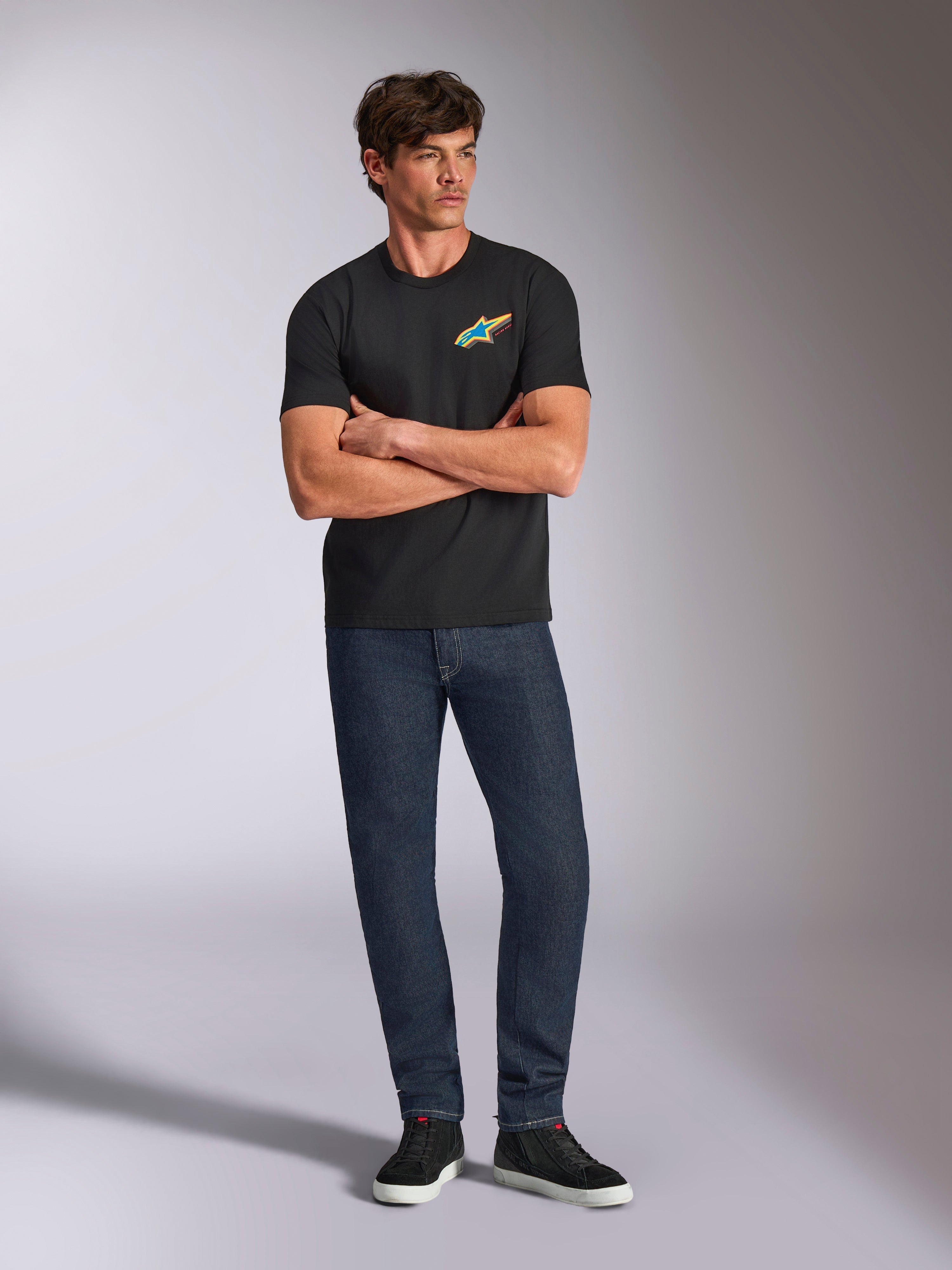 Alpinestars Angler CSF Tee, maglietta a maniche corte, nera, vivace logo Astars multicolore sul petto, indossata da un modello con le braccia incrociate, jeans in denim scuro e sneakers nere, streetwear casual motorsport