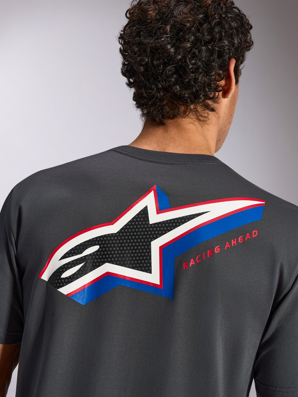 Alpinestars Angler CSF Tee, maglietta a maniche corte, grigio scuro, vista posteriore su modello con grande logo a stella Alpinestars multicolore e riempimento traforato, scritta 'RACING AHEAD', abbigliamento casual motorsport streetwear
