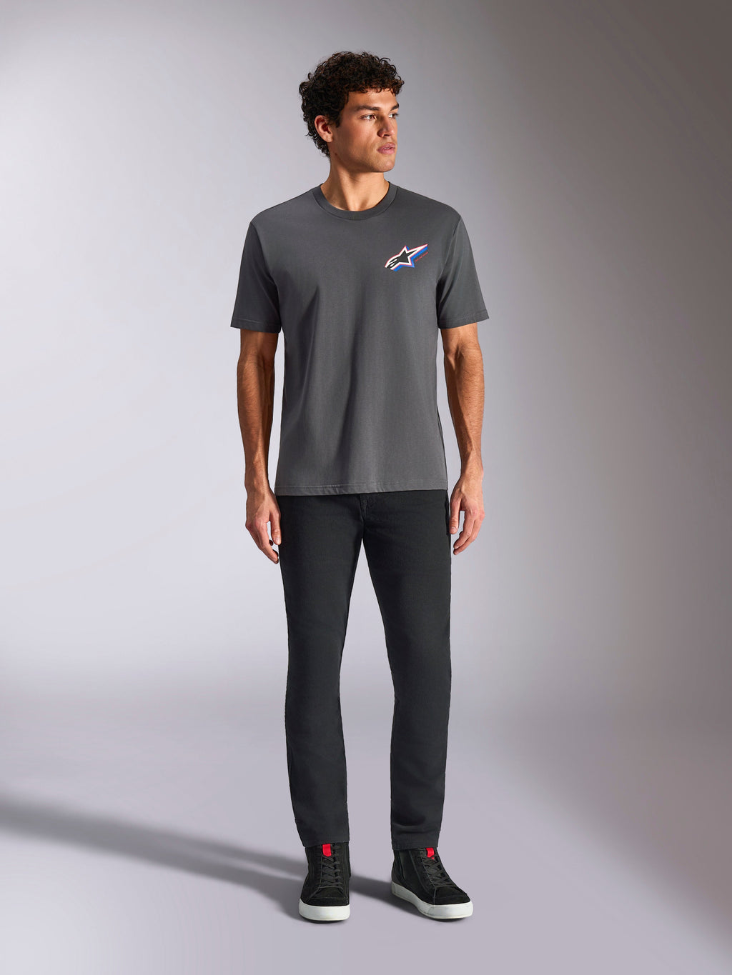 Alpinestars Angler CSF Tee, maglietta a maniche corte, antracite, con vivace logo Astars multicolore sul petto, indossata da un modello con pantaloni neri e sneakers, streetwear casual motorsport
