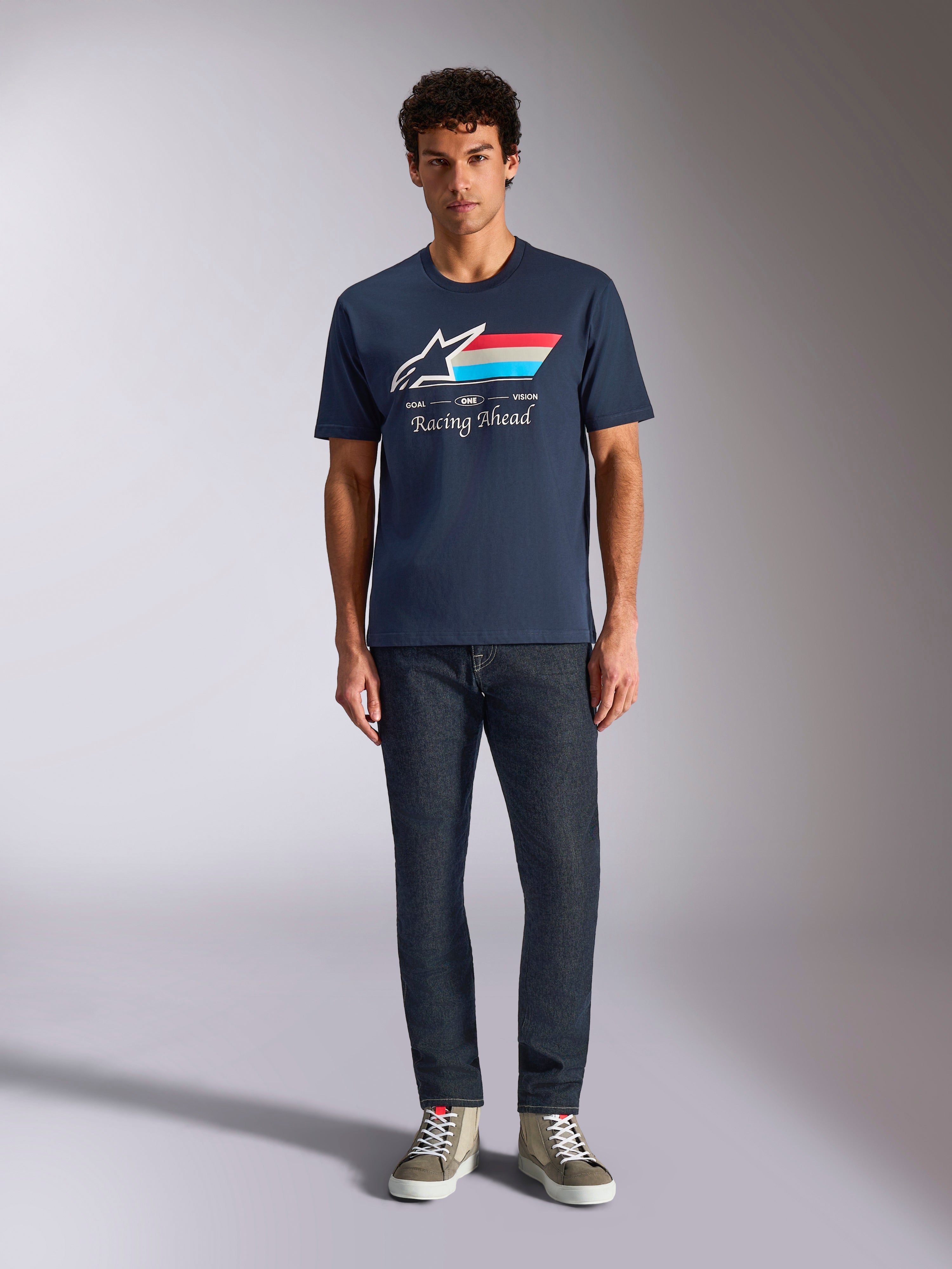 Alpinestars Trifecta CSF Tee, t-shirt a maniche corte, blu navy, logo Astars bianco con strisce rosse, bianche e azzurre e scritta 'Racing Ahead', indossata con jeans scuri e sneakers verde oliva, abbigliamento casual motorsport streetwear