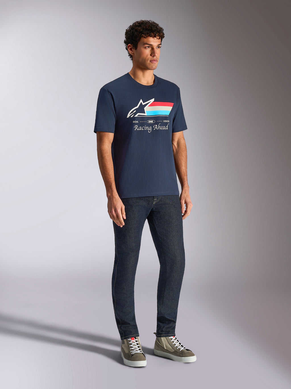 Alpinestars Trifecta CSF Tee, t-shirt a maniche corte, blu navy, con logo Astars a strisce tricolore e testo Racing Ahead, indossata da un modello con jeans scuri e sneakers, streetwear casual ispirato al motorsport