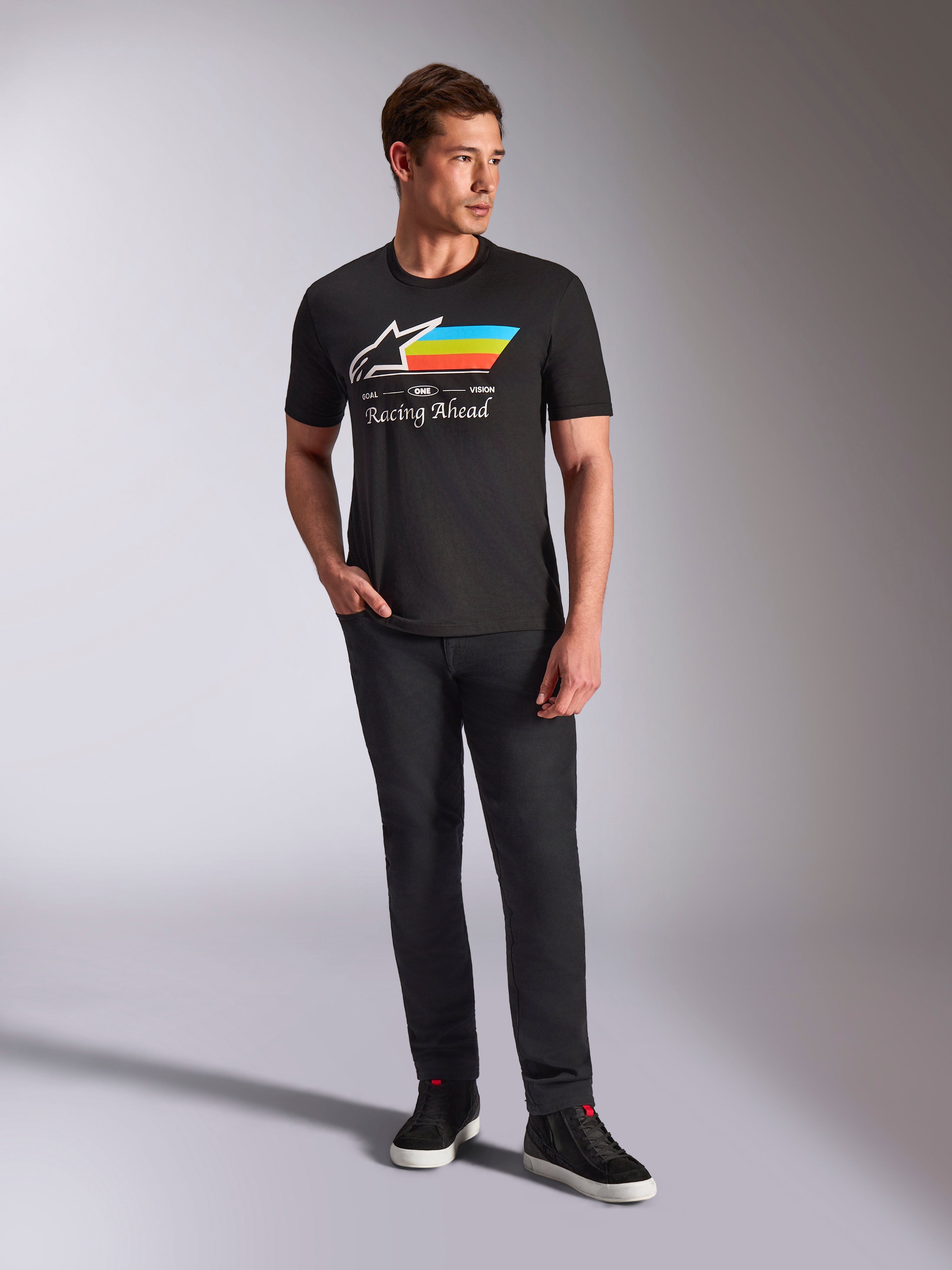 Alpinestars Trifecta CSF Tee, t-shirt a maniche corte, nero, con logo Astars bianco con strisce ciano, gialle e arancioni e scritta 'Racing Ahead', indossata da un modello con pantaloni neri e sneakers per un look motorsport streetwear casual