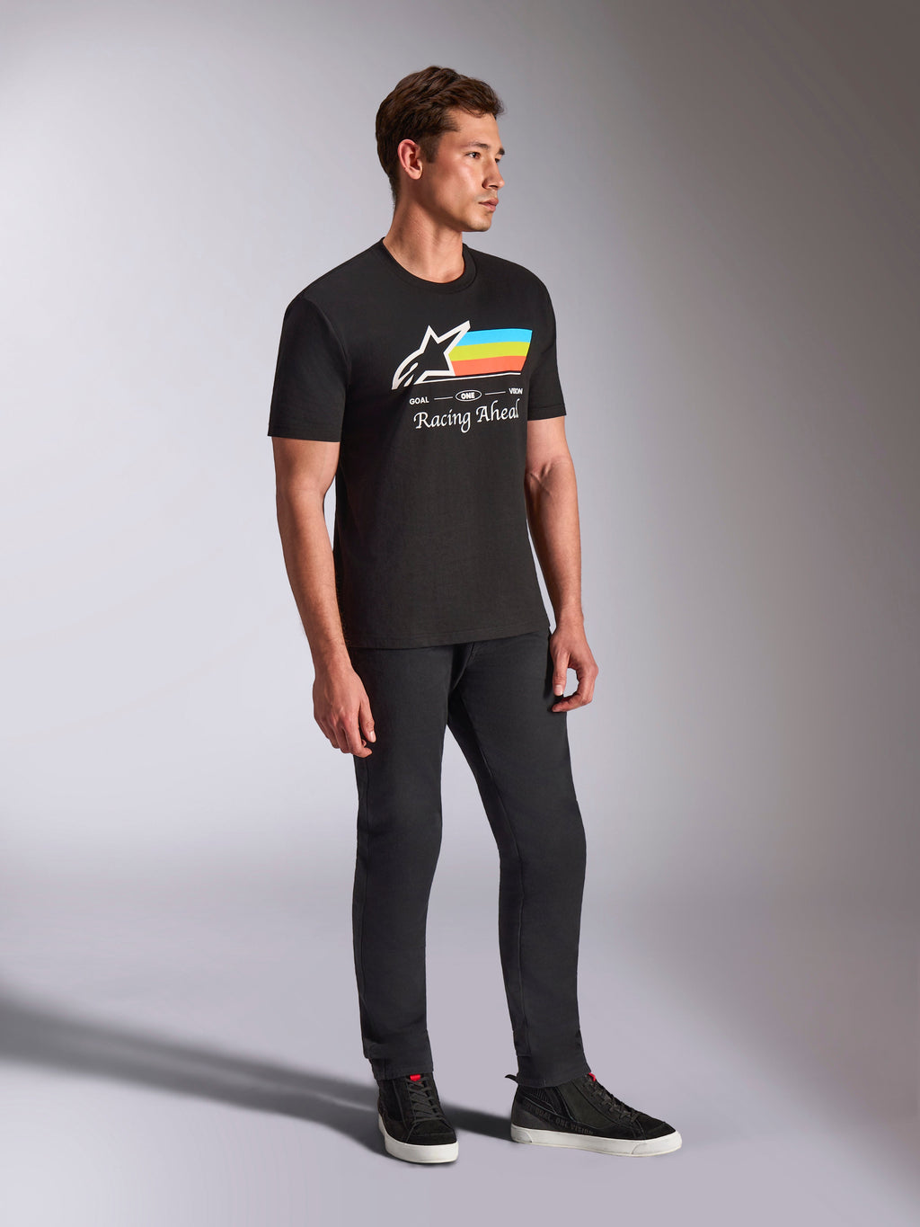 Alpinestars Trifecta CSF Tee, maglietta a maniche corte nera, logo Astars bianco con strisce blu, gialle e arancioni e grafica 'Racing Ahead', indossata da un modello con pantaloni neri e sneakers, abbigliamento lifestyle casual ispirato al motorsport