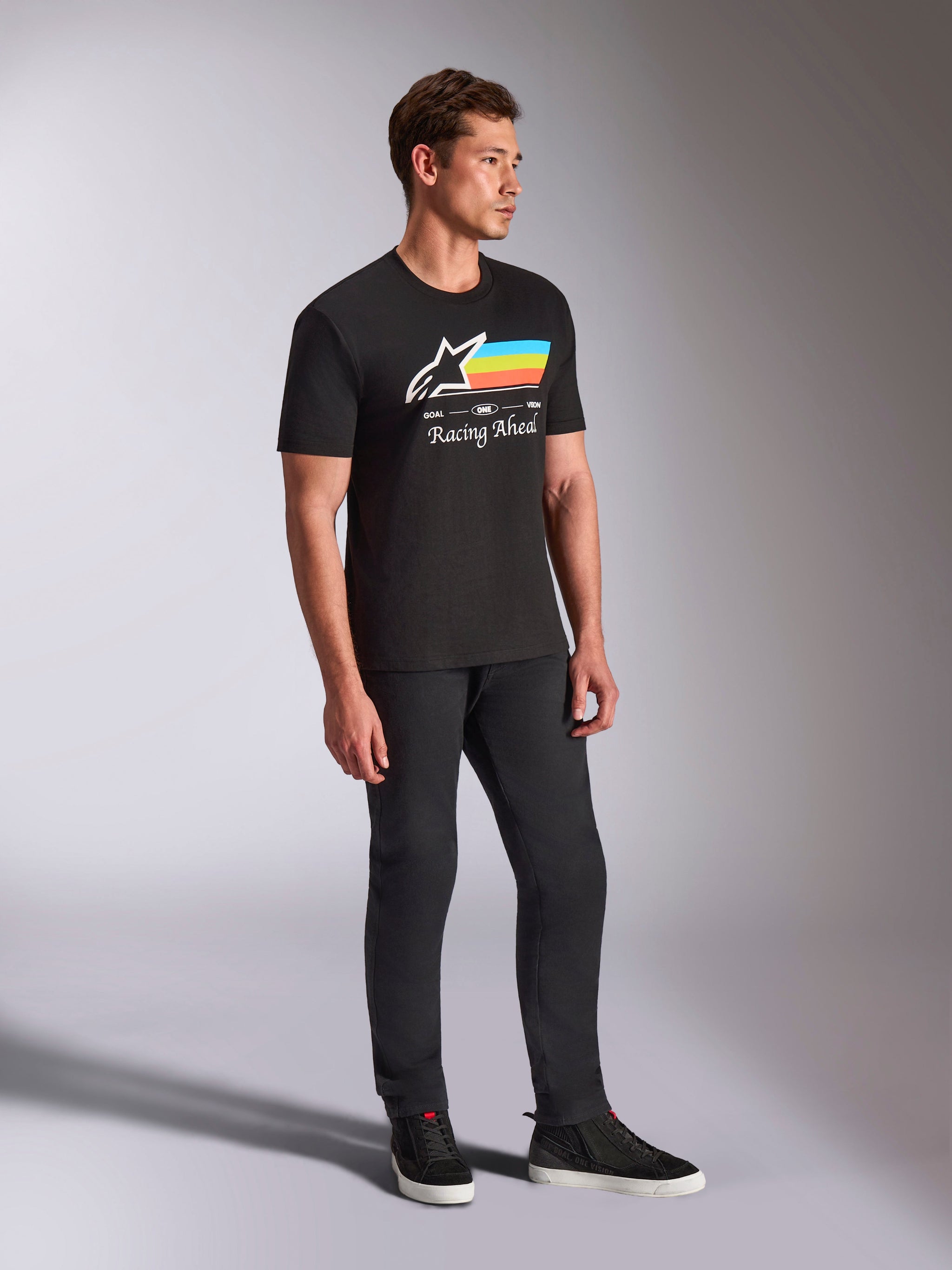 Alpinestars Trifecta CSF Tee, maglietta a maniche corte nera, logo Astars bianco con strisce blu, gialle e arancioni e grafica 'Racing Ahead', indossata da un modello con pantaloni neri e sneakers, abbigliamento lifestyle casual ispirato al motorsport