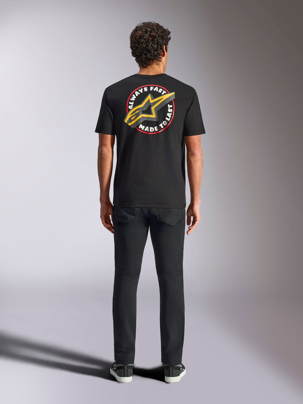 T-shirt Alpinestars Aorbit CSF, maglietta a maniche corte, nero, indossata da un modello vista dal retro con pantaloni neri, con grande grafica 'Always Fast Made to Last' e logo Astars giallo, streetwear casual a tema motorsport