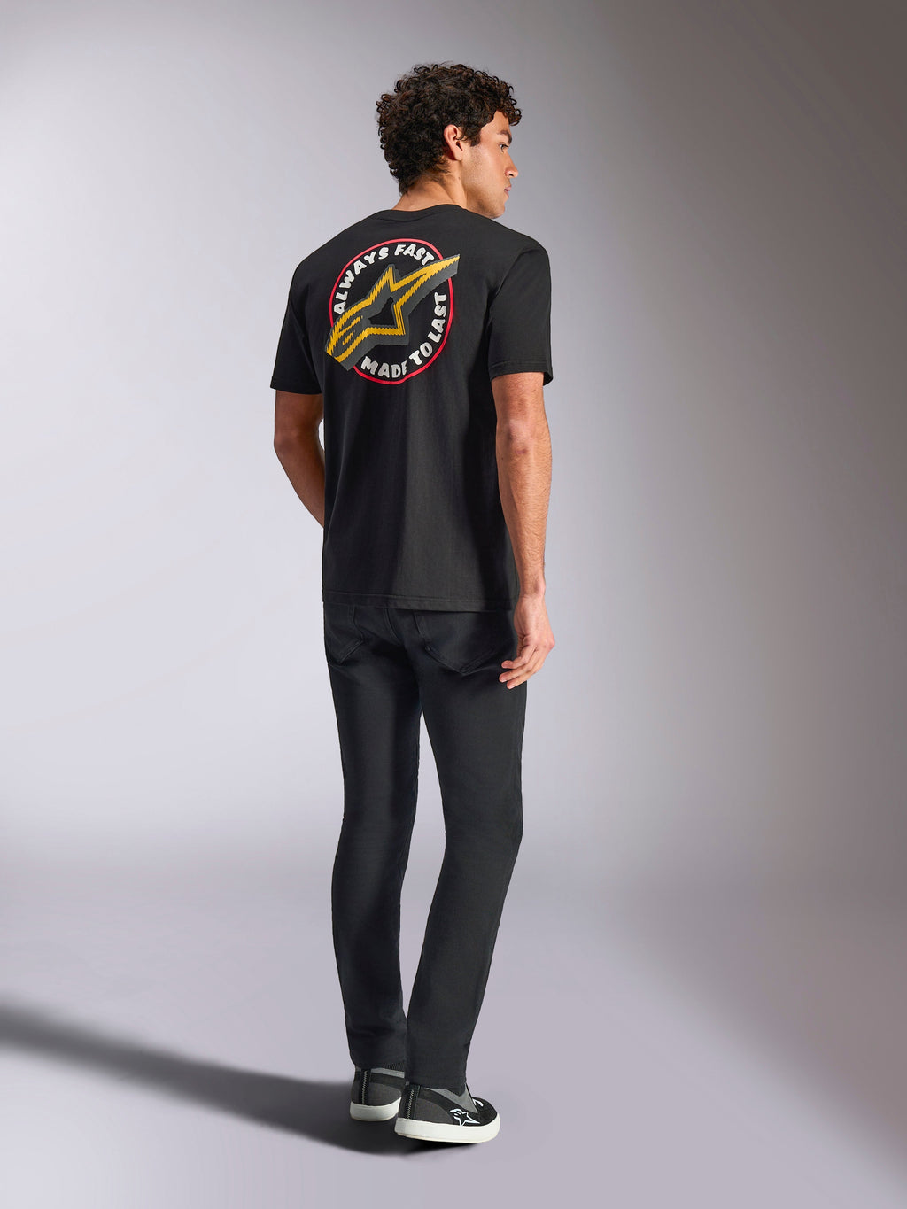 Alpinestars Aorbit CSF Tee, t-shirt a maniche corte, nero, vista posteriore con grande logo Astars giallo e grafica circolare 'Always Fast Made to Last', indossata con jeans neri e sneakers casual, stile streetwear ispirato al motorsport