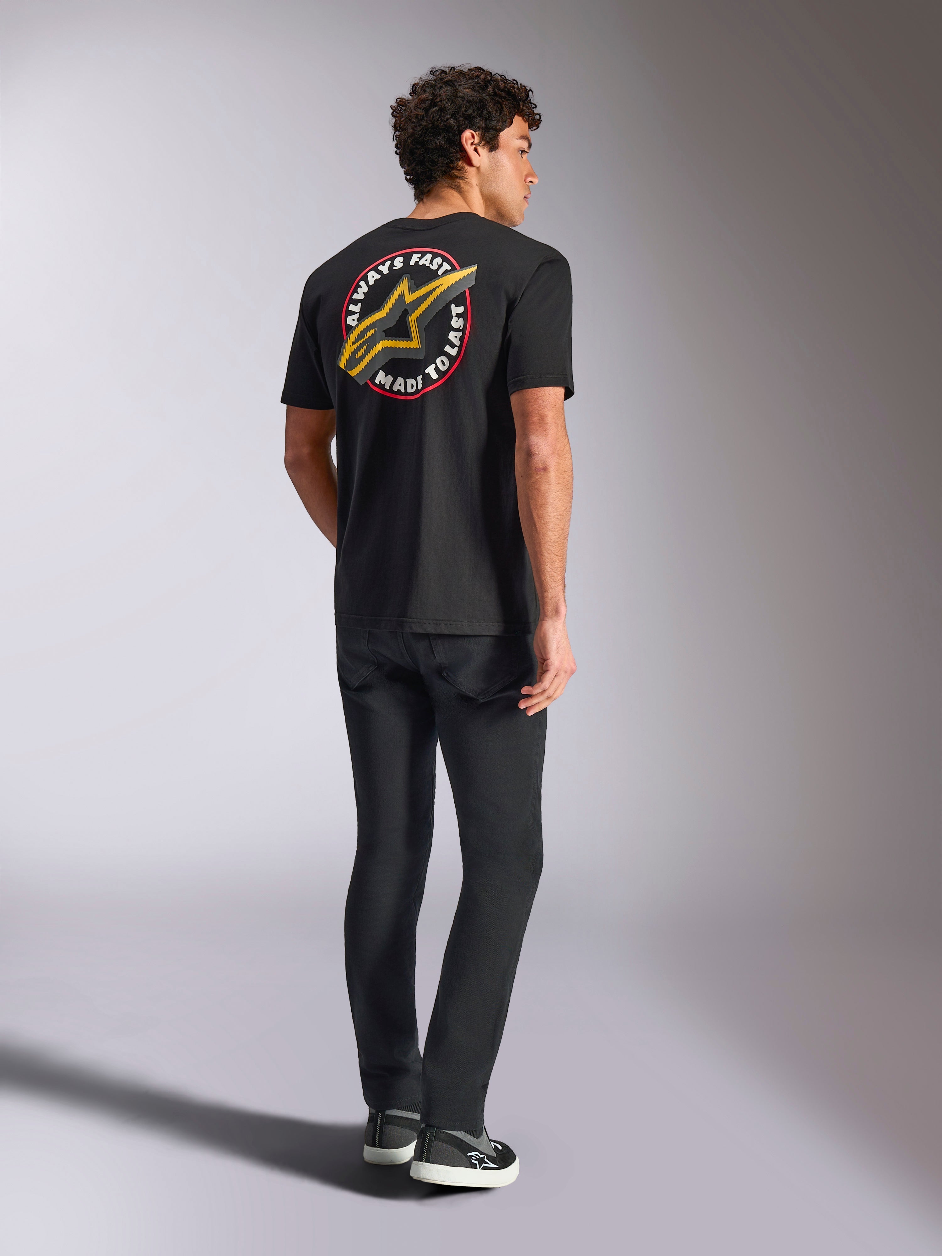 Alpinestars Aorbit CSF Tee, t-shirt a maniche corte, nero, vista posteriore con grande logo Astars giallo e grafica circolare 'Always Fast Made to Last', indossata con jeans neri e sneakers casual, stile streetwear ispirato al motorsport