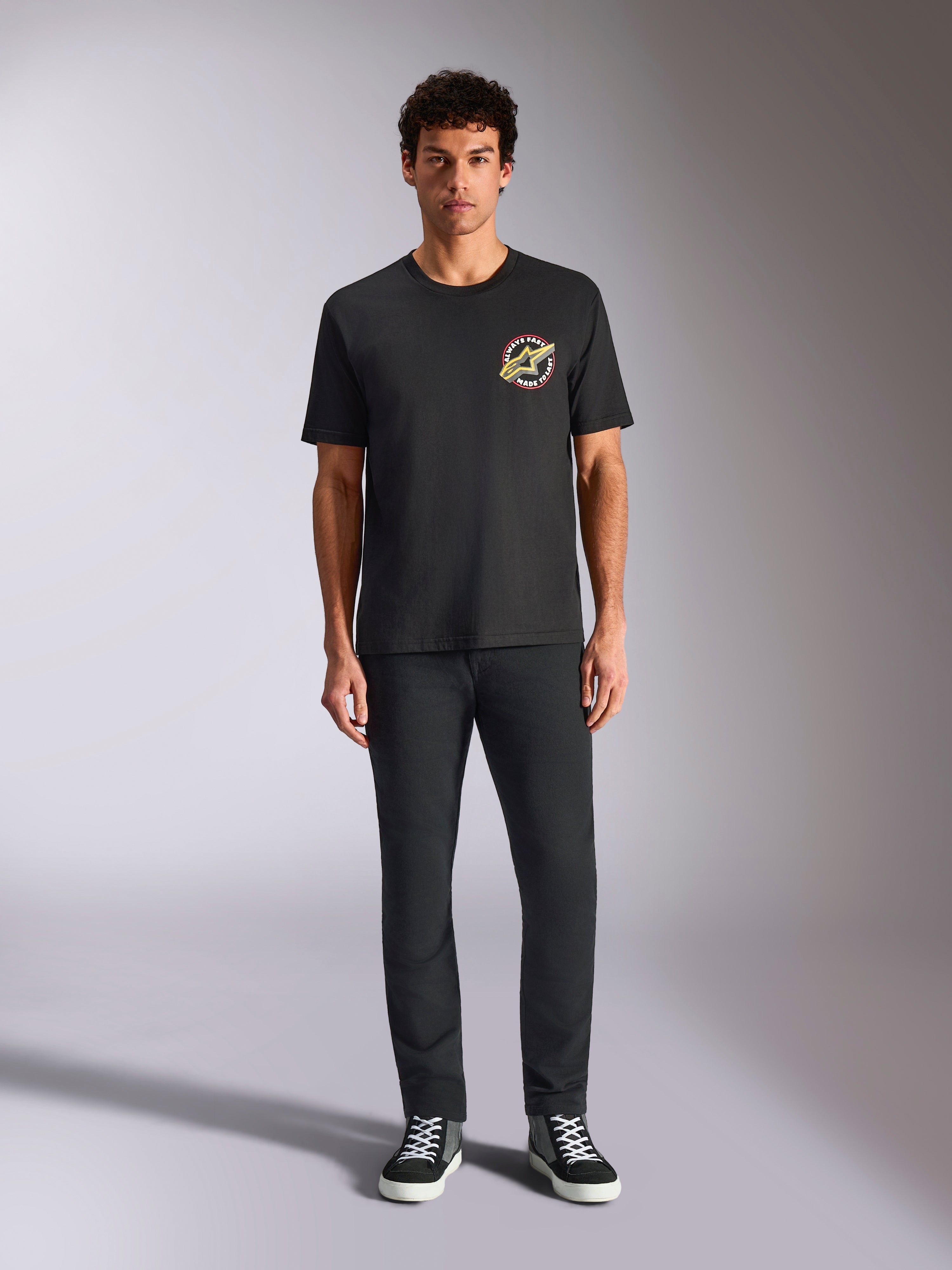 Alpinestars Aorbit CSF Tee, t-shirt a maniche corte, nera, logo circolare con scritta 'Always Fast Made to Last' sul petto, indossata da un modello con pantaloni neri e sneakers, streetwear casual ispirato al motorsport