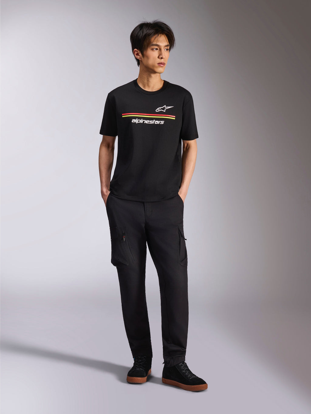 Modello che indossa la Alpinestars Lanes CSF Tee, t-shirt a maniche corte nera, con strisce rosse e gialle e loghi Astars e Alpinestars, abbinata a pantaloni cargo neri e sneakers, streetwear casual motorsport