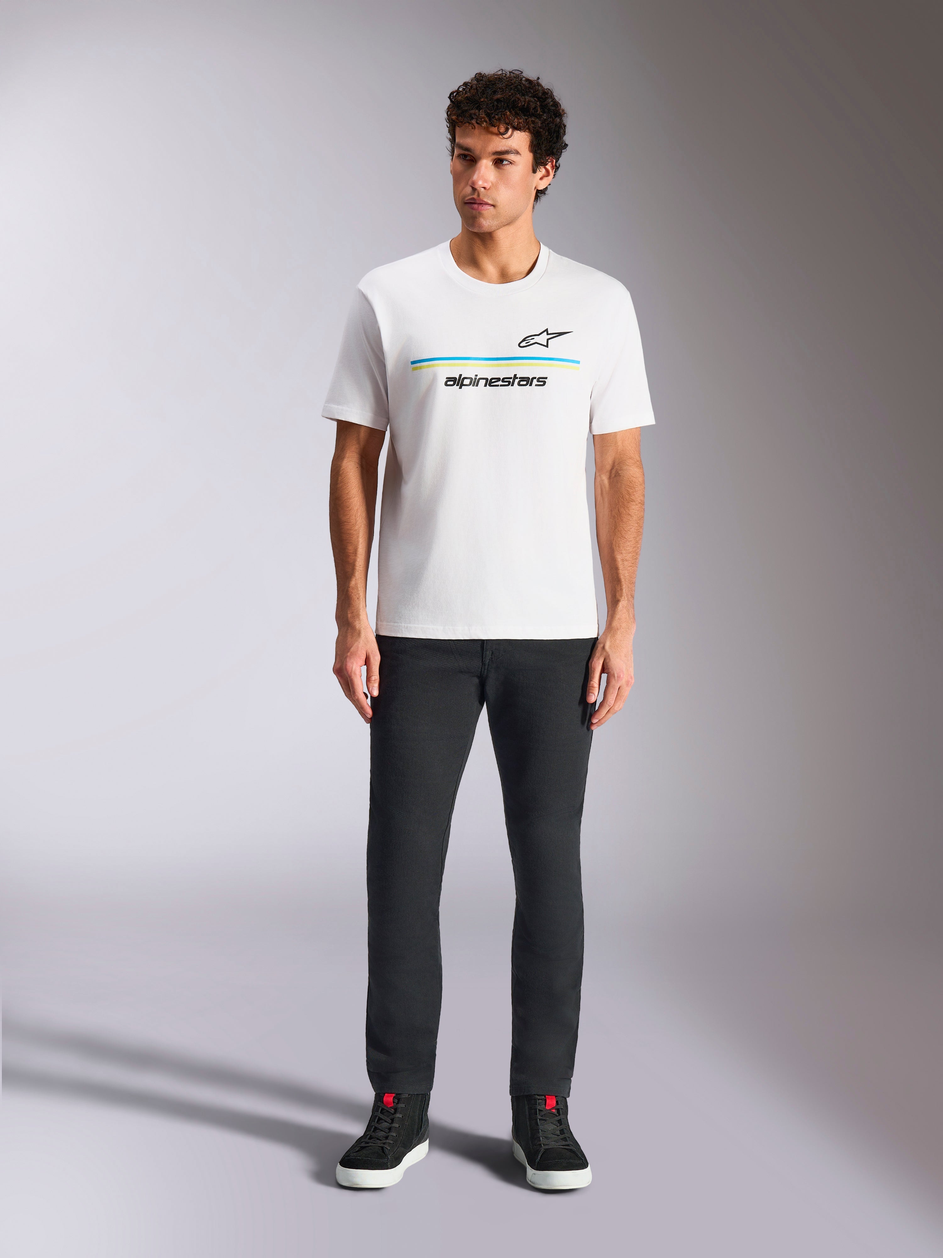 Alpinestars Lanes CSF Tee, t-shirt a maniche corte, bianca, grafica con logo a strisce blu e gialle, indossata da un modello con pantaloni neri e sneakers, streetwear casual ispirato al motorsport