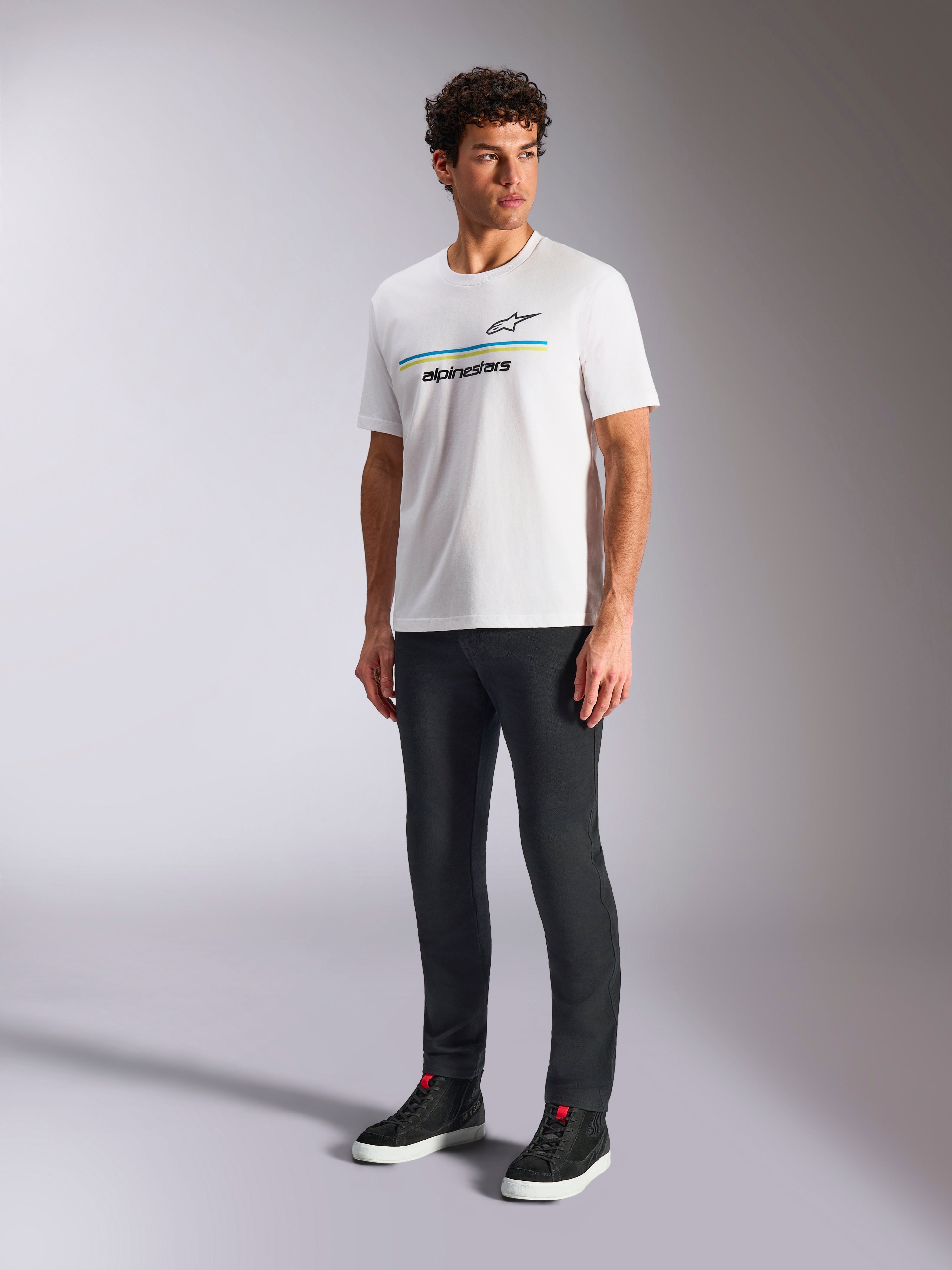 T-shirt Alpinestars Lanes CSF, maglietta bianca a maniche corte, indossata da un modello con pantaloni neri slim-fit e sneakers alte nere, con strisce orizzontali blu e gialle e logo Alpinestars sul petto, streetwear casual ispirato al motorsport