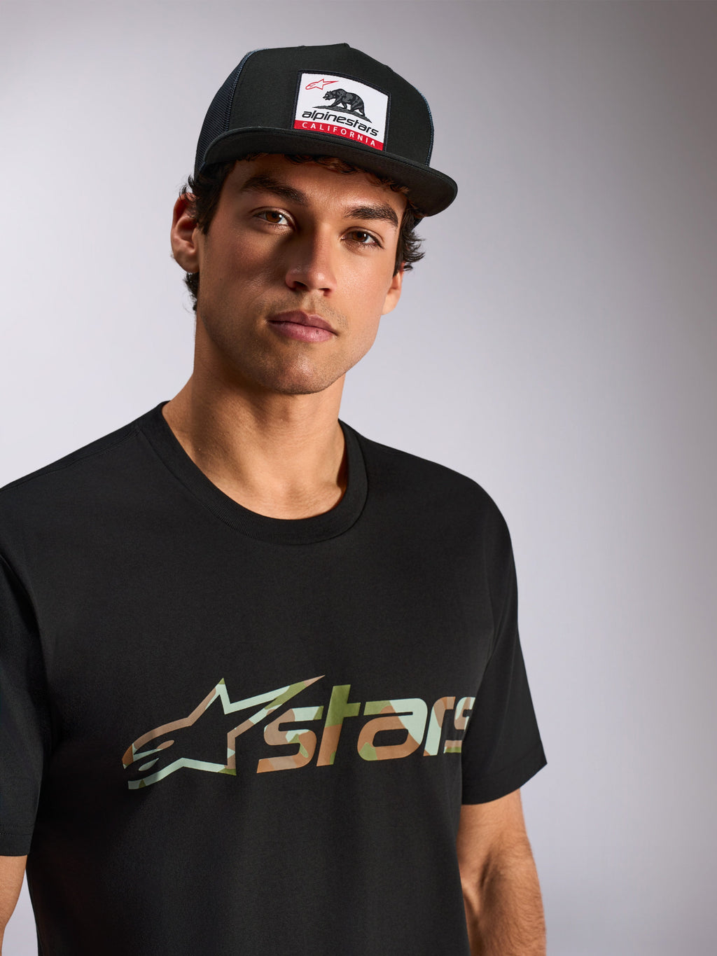 Alpinestars Verdure Camo CSF Tee, t-shirt a maniche corte, nera, logo Astars con riempimento camo, indossata da un modello con cappello trucker Alpinestars California nero, abbigliamento casual motorsport streetwear