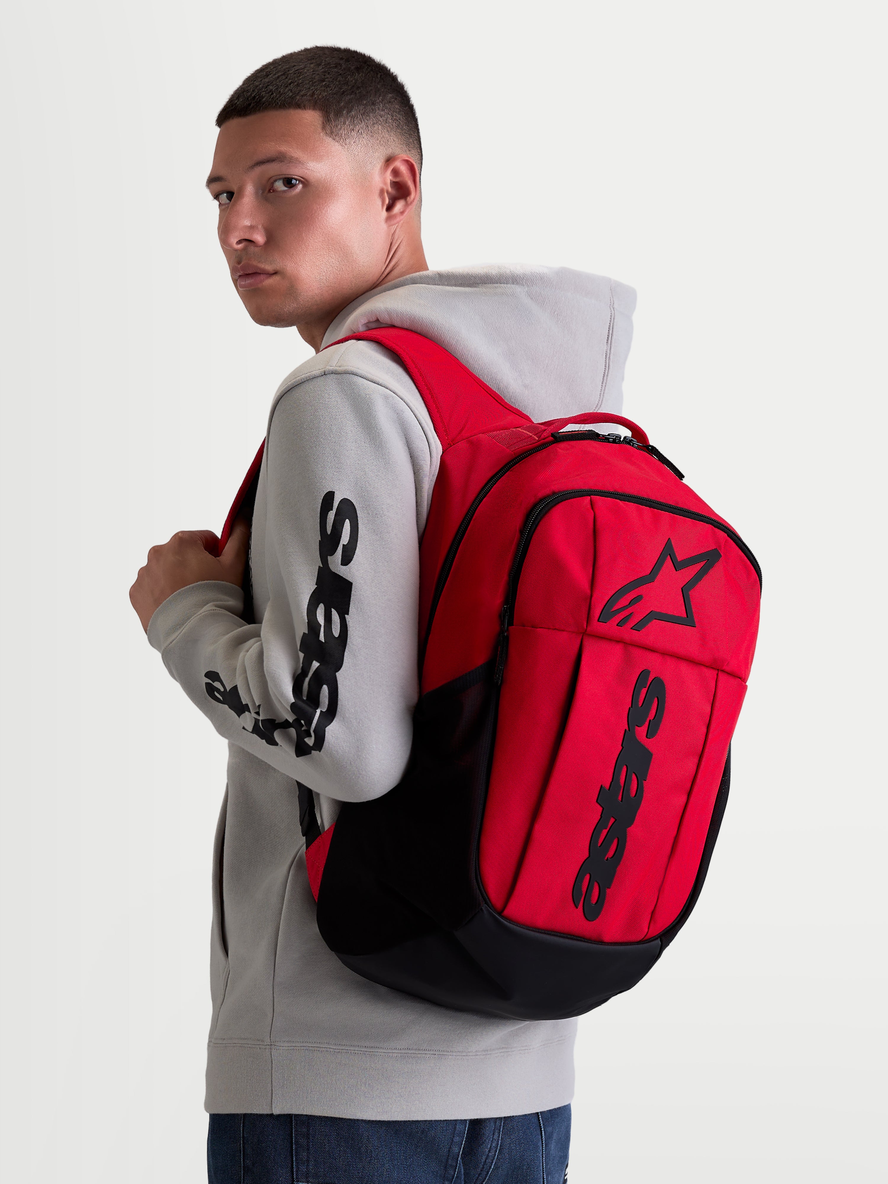 Alpinestars Zaino GFX Boss, zaino sportivo urban da 20L, rosso e nero, indossato da un modello con felpa grigia. Caratterizzato da una struttura in poliestere 600D, scomparto interno per laptop e grafica Astars in silicone in rilievo. Ideale per l'uso quotidiano.