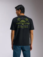 Alpinestars Order CSF Tee, t-shirt a maniche corte, nero, vista posteriore, grafica verde oliva con palme e logo del casco, testo 'ONE GOAL. ONE VISION. ALPINESTARS', abbinata a jeans blu, streetwear casual motorsport