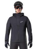 Giacca Alpinestars A-Dura Elite Waterproof, nero, giacca da mountain bike, design a collo alto con cappuccio, cerniera frontale con patta protettiva sul petto e logo bianco, indossata con casco nero e guanti da ciclismo, protezione tecnica contro le intemperie per il trail riding