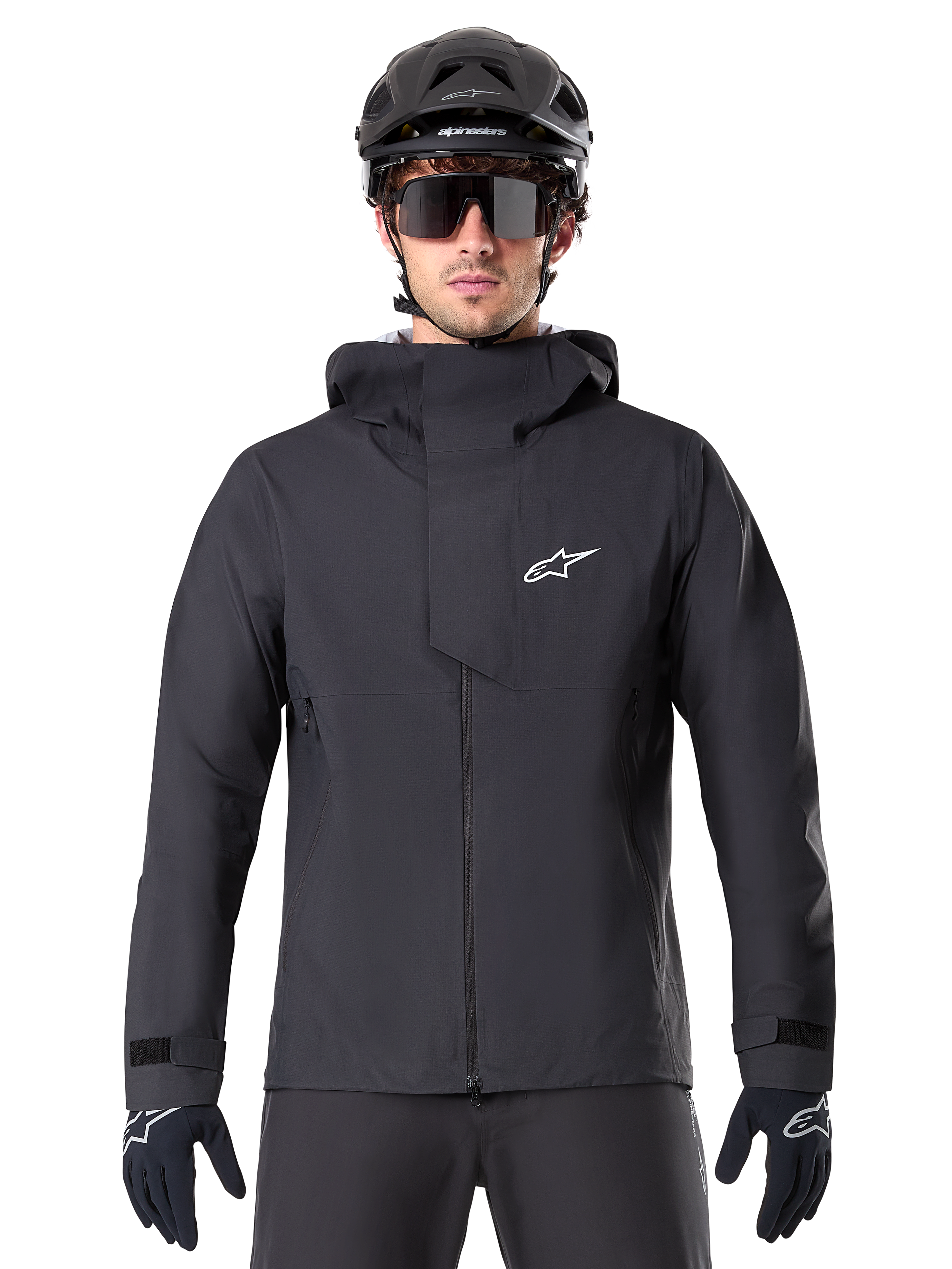 Giacca Alpinestars A-Dura Elite Waterproof, nero, giacca da mountain bike, design a collo alto con cappuccio, cerniera frontale con patta protettiva sul petto e logo bianco, indossata con casco nero e guanti da ciclismo, protezione tecnica contro le intemperie per il trail riding