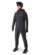 Giacca impermeabile Alpinestars A-Dura Elite in nero, indossata da un pilota uomo con pantaloni da MTB, guanti e casco neri coordinati, design tecnico impermeabile, logo Astars bianco sul petto e collo alto protettivo.