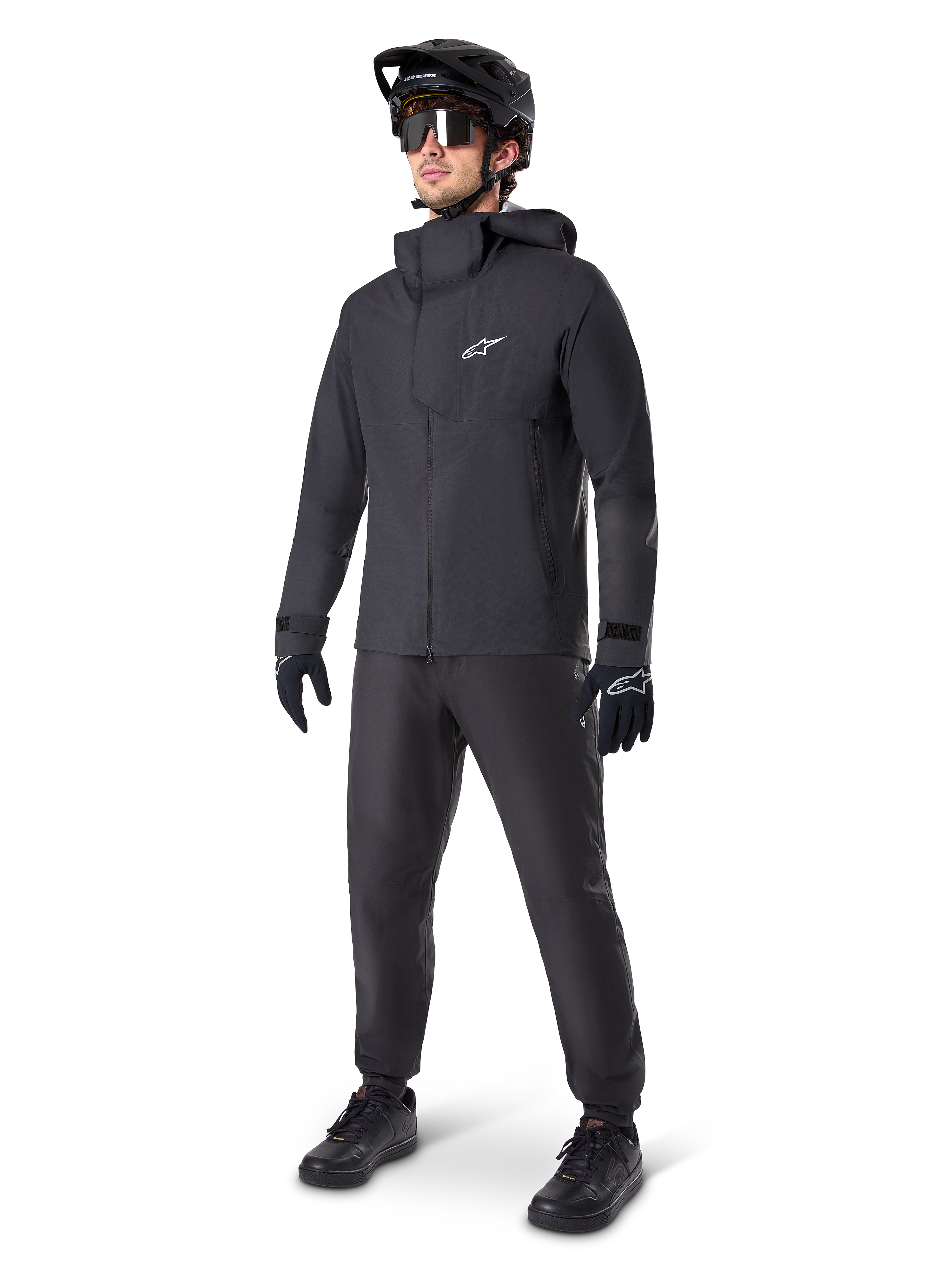 Giacca impermeabile Alpinestars A-Dura Elite in nero, indossata da un pilota uomo con pantaloni da MTB, guanti e casco neri coordinati, design tecnico impermeabile, logo Astars bianco sul petto e collo alto protettivo.