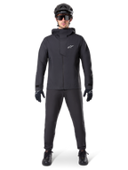 Giacca impermeabile Alpinestars A-Dura Elite, colore nero, indossata con pantaloni da ciclismo e guanti coordinati, casco e occhiali da sole neri, con logo Astars bianco sul petto, design tecnico ad alte prestazioni per mountain bike