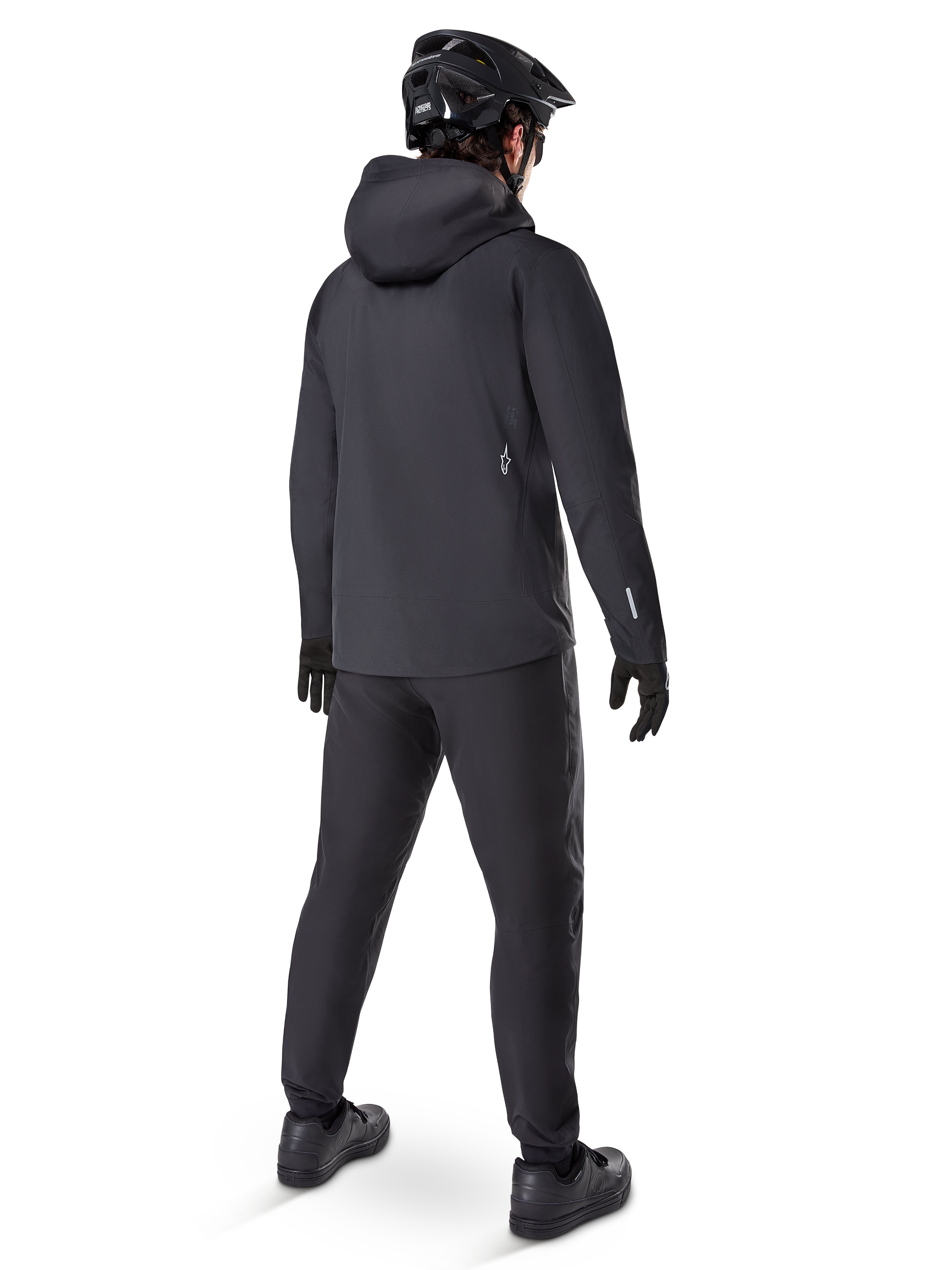 Giacca impermeabile Alpinestars A-Dura Elite, Nero, vista posteriore con cappuccio sopra il casco e orlo posteriore più lungo, abbinata a pantaloni MTB neri coordinati, guanti e casco per una protezione completa sui sentieri in ogni condizione atmosferica.