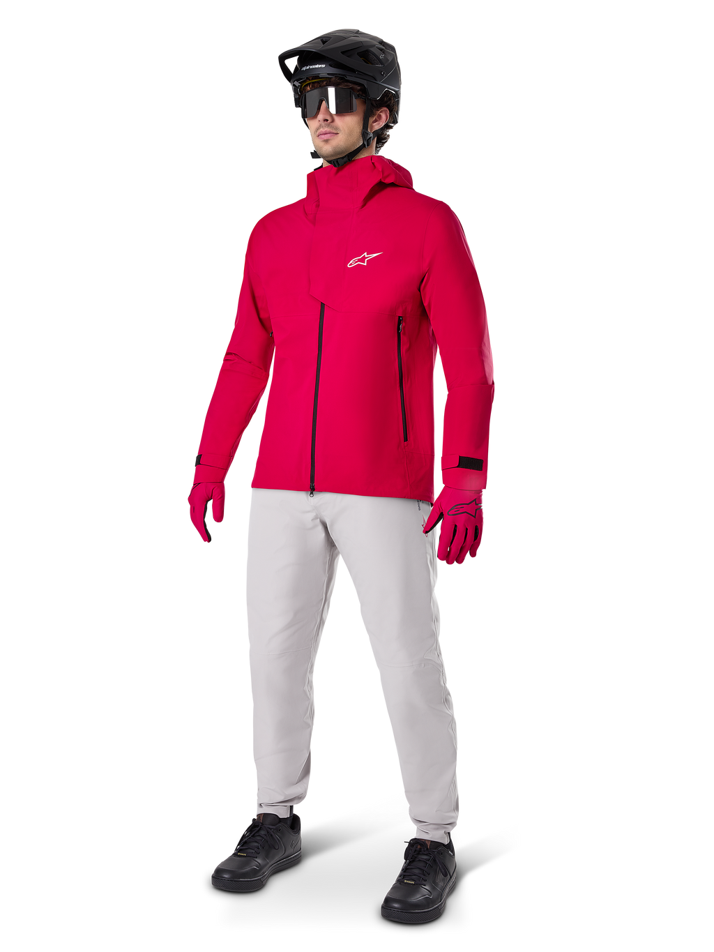 Ciclista con giacca impermeabile Alpinestars A-Dura Elite in Red Fluo, pantaloni tecnici grigio chiaro, guanti rossi, casco da MTB nero con maschera e scarpe nere, abbigliamento completo ad alte prestazioni per mountain bike