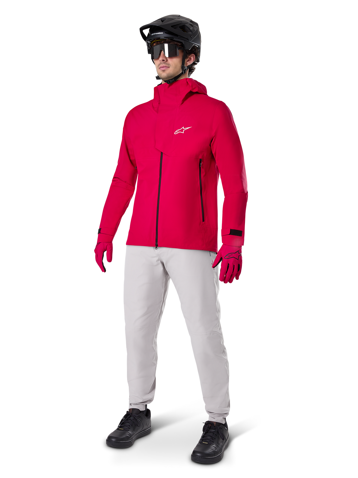 Ciclista con giacca impermeabile Alpinestars A-Dura Elite in Red Fluo, pantaloni tecnici grigio chiaro, guanti rossi, casco da MTB nero con maschera e scarpe nere, abbigliamento completo ad alte prestazioni per mountain bike
