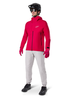Ciclista con giacca impermeabile Alpinestars A-Dura Elite in Red Fluo, pantaloni tecnici grigio chiaro, guanti rossi, casco da MTB nero con maschera e scarpe nere, abbigliamento completo ad alte prestazioni per mountain bike