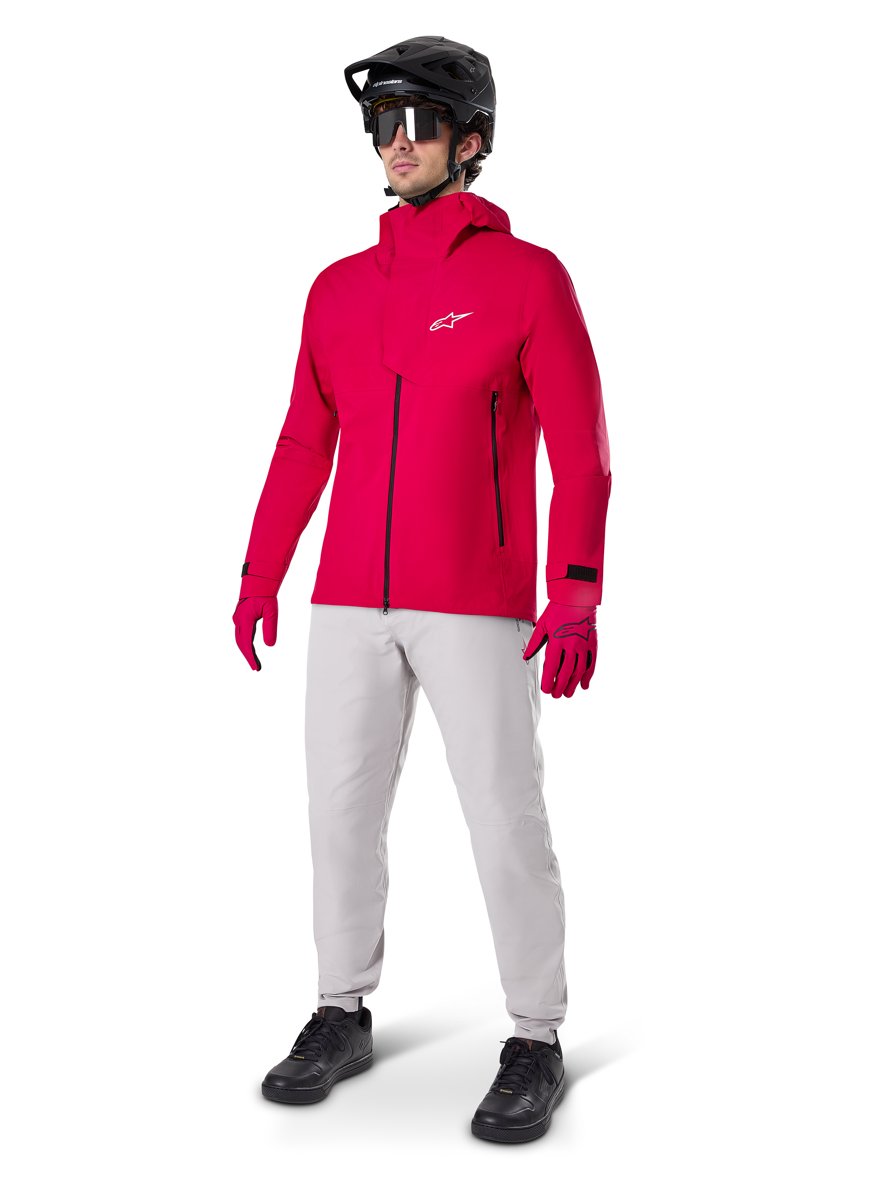 Ciclista con giacca impermeabile Alpinestars A-Dura Elite in Red Fluo, pantaloni tecnici grigio chiaro, guanti rossi, casco da MTB nero con maschera e scarpe nere, abbigliamento completo ad alte prestazioni per mountain bike