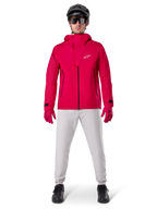 Giacca impermeabile Alpinestars A-Dura Elite, Rosso Fluo, giacca tecnica da mountain bike indossata con un casco da MTB nero, occhiali da sole, guanti rossi coordinati e pantaloni grigio chiaro, vista a figura intera dell'abbigliamento da ciclismo per tutte le condizioni atmosferiche