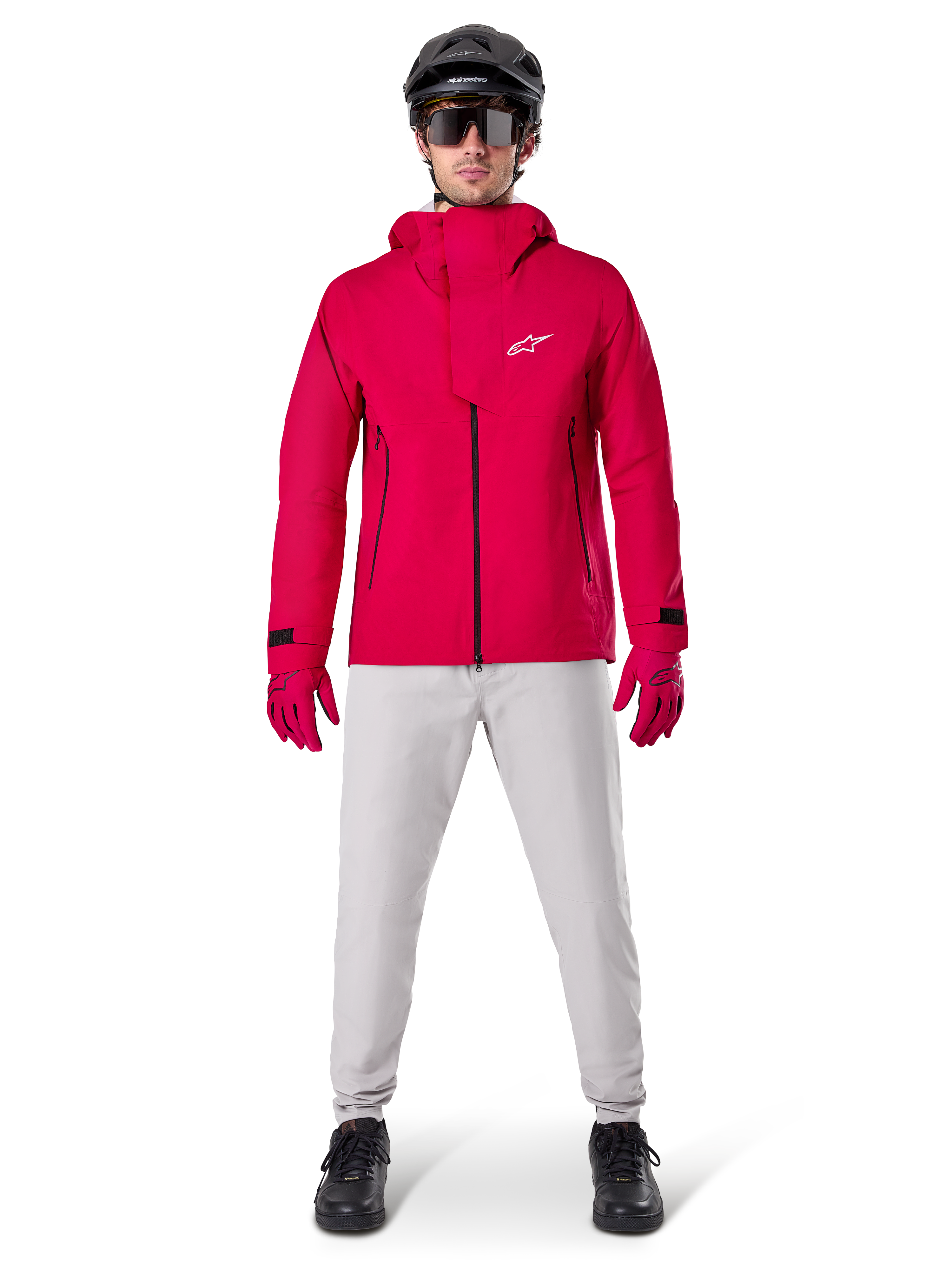 Giacca impermeabile Alpinestars A-Dura Elite, Rosso Fluo, giacca tecnica da mountain bike indossata con un casco da MTB nero, occhiali da sole, guanti rossi coordinati e pantaloni grigio chiaro, vista a figura intera dell'abbigliamento da ciclismo per tutte le condizioni atmosferiche