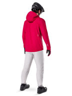 Alpinestars Giacca Impermeabile A-Dura Elite, Rosso Fluo, vista posteriore a tre quarti, indossata con pantaloni grigio chiaro e casco MTB nero, dotata di cappuccio e dettagli riflettenti, giacca antipioggia da ciclismo ad alte prestazioni per trail riding