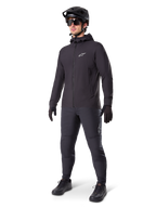 Giacca termica Alpinestars A-Dura in nero, indossata da un ciclista con equipaggiamento completo da mountain bike tra cui casco nero, occhiali da sole e pantaloni da ciclismo, giacca con cappuccio leggera e isolata per prestazioni ciclistiche in climi freddi