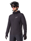 Alpinestars A-Dura Thermal Jacket, giacca da ciclismo nera, design con cappuccio e zip frontale, logo a stella bianco sul petto, indossata con casco da mountain bike e guanti neri, abbigliamento sportivo per il trail riding in climi freddi